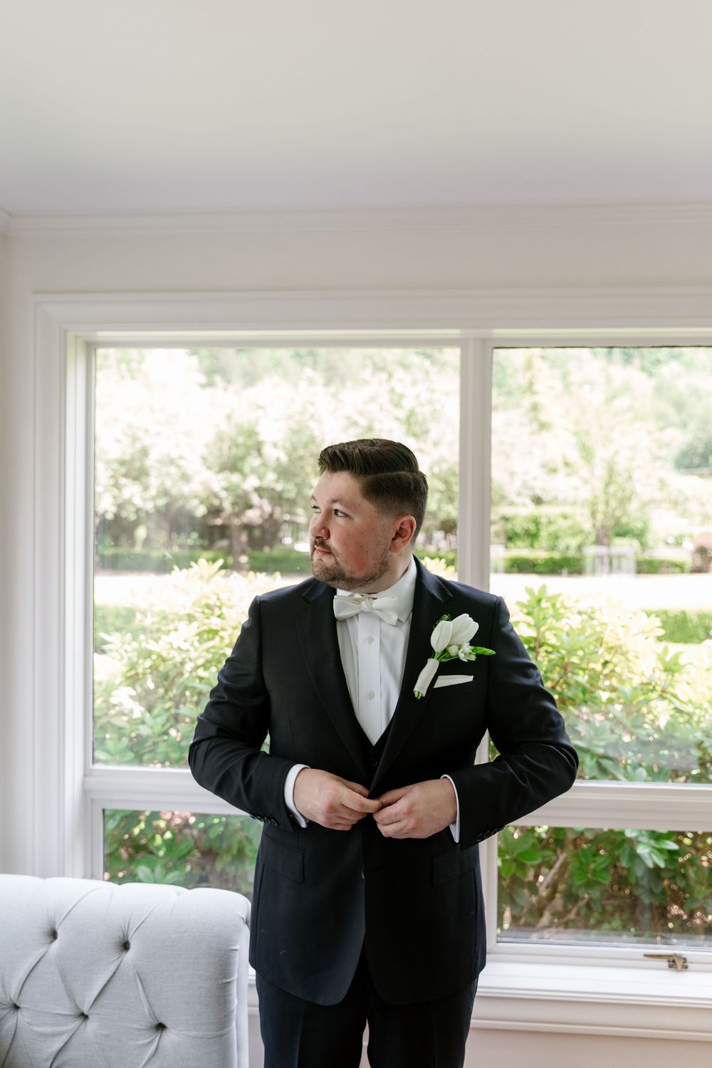 A person in a black tuxedo adjusts their bowtie while standing near bright windows in an elegant room.