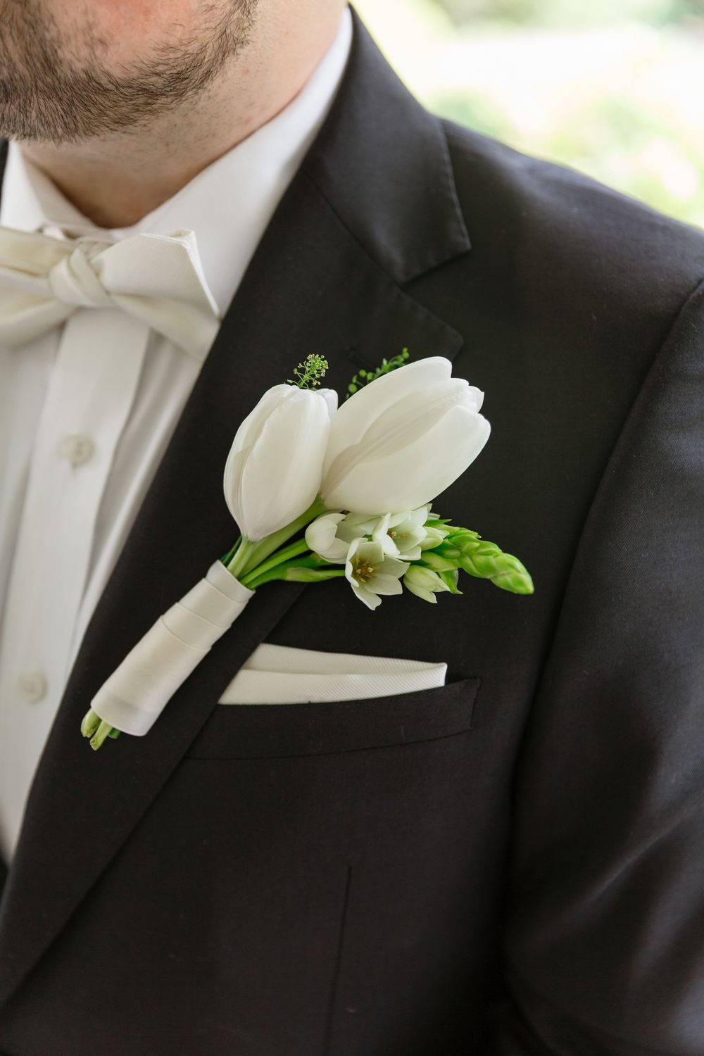Elegant white tulip and lily boutonniere pinned to a black tuxedo lapel with matching white bow tie.
