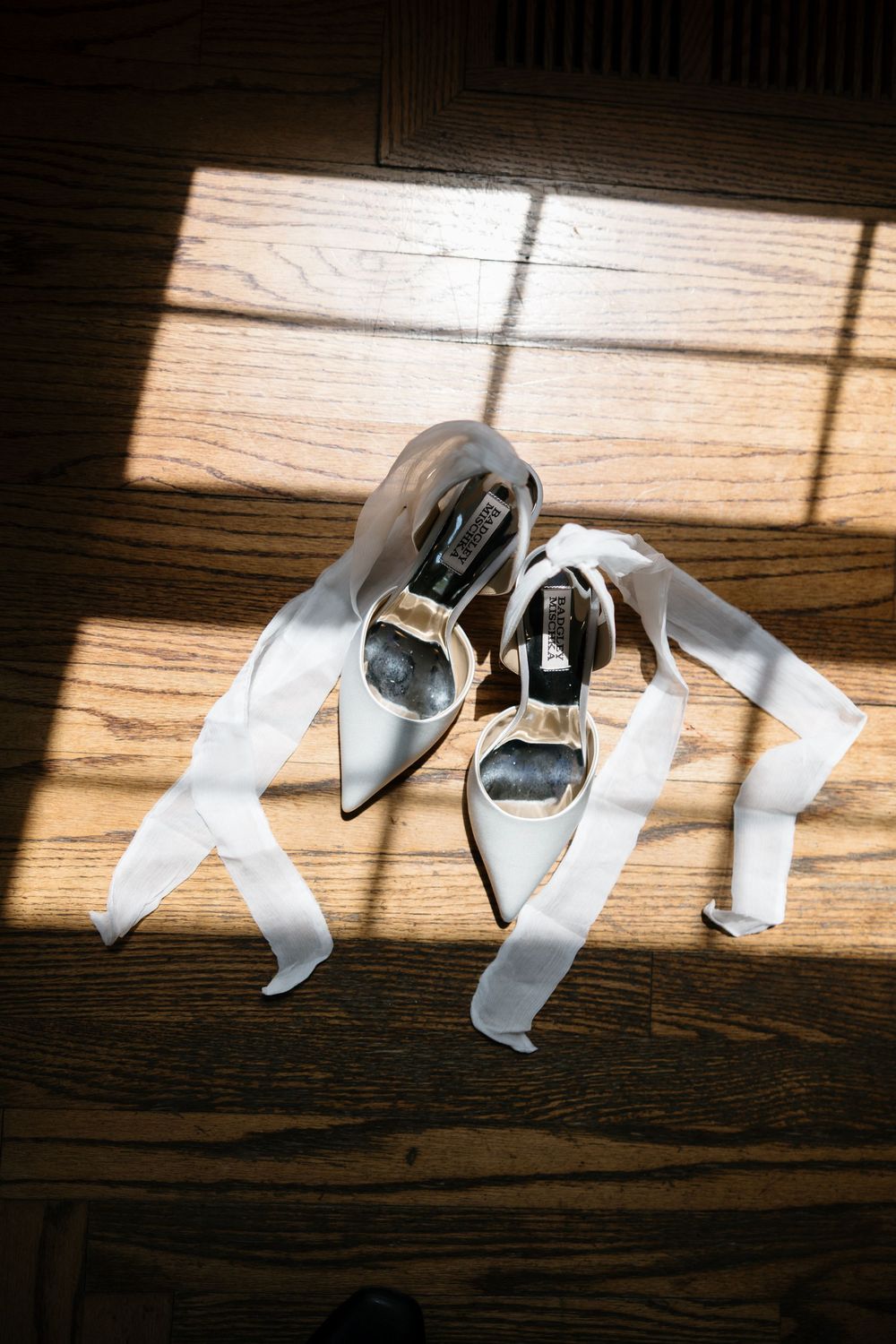 Silver bridal shoes and white ribbons arranged artistically on a wooden floor in sunlight.