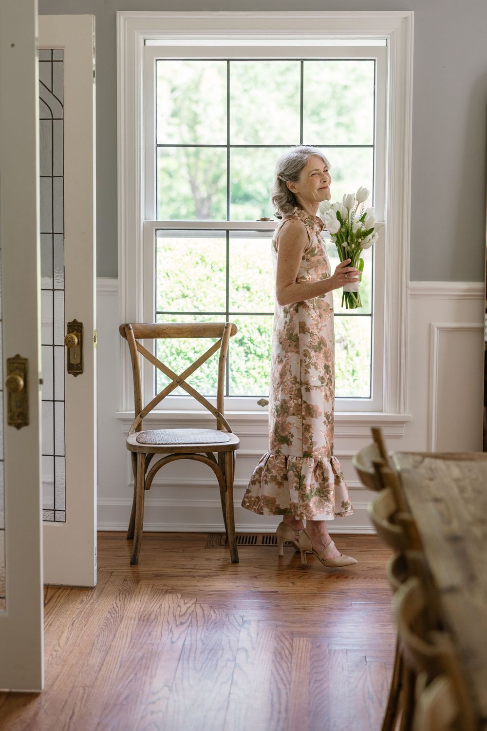 A person in a floral dress stands by a bright window holding a bouquet of white tulips in an elegant interior setting.
