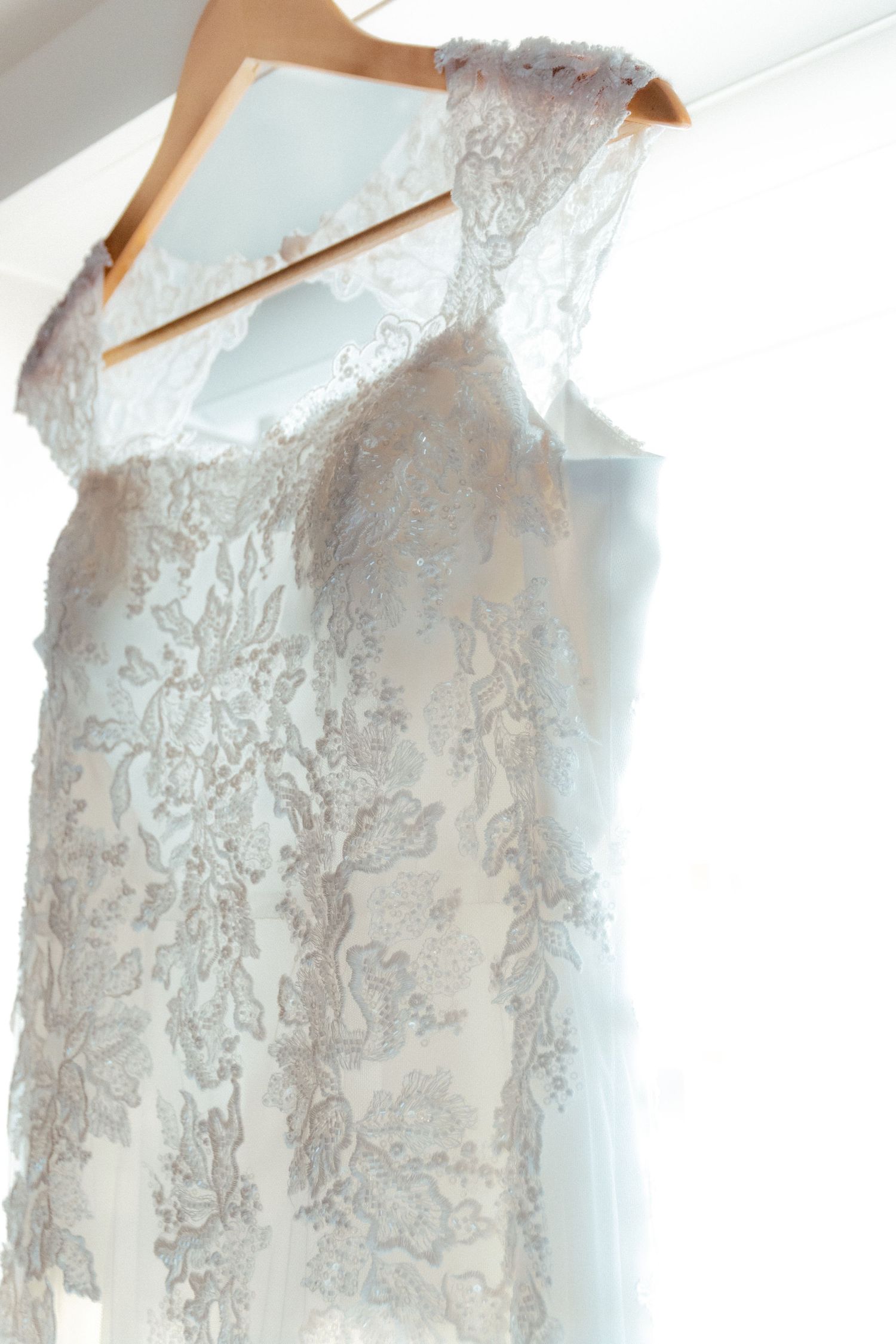 A delicate white lace wedding dress with intricate floral patterns hanging on a wooden hanger.