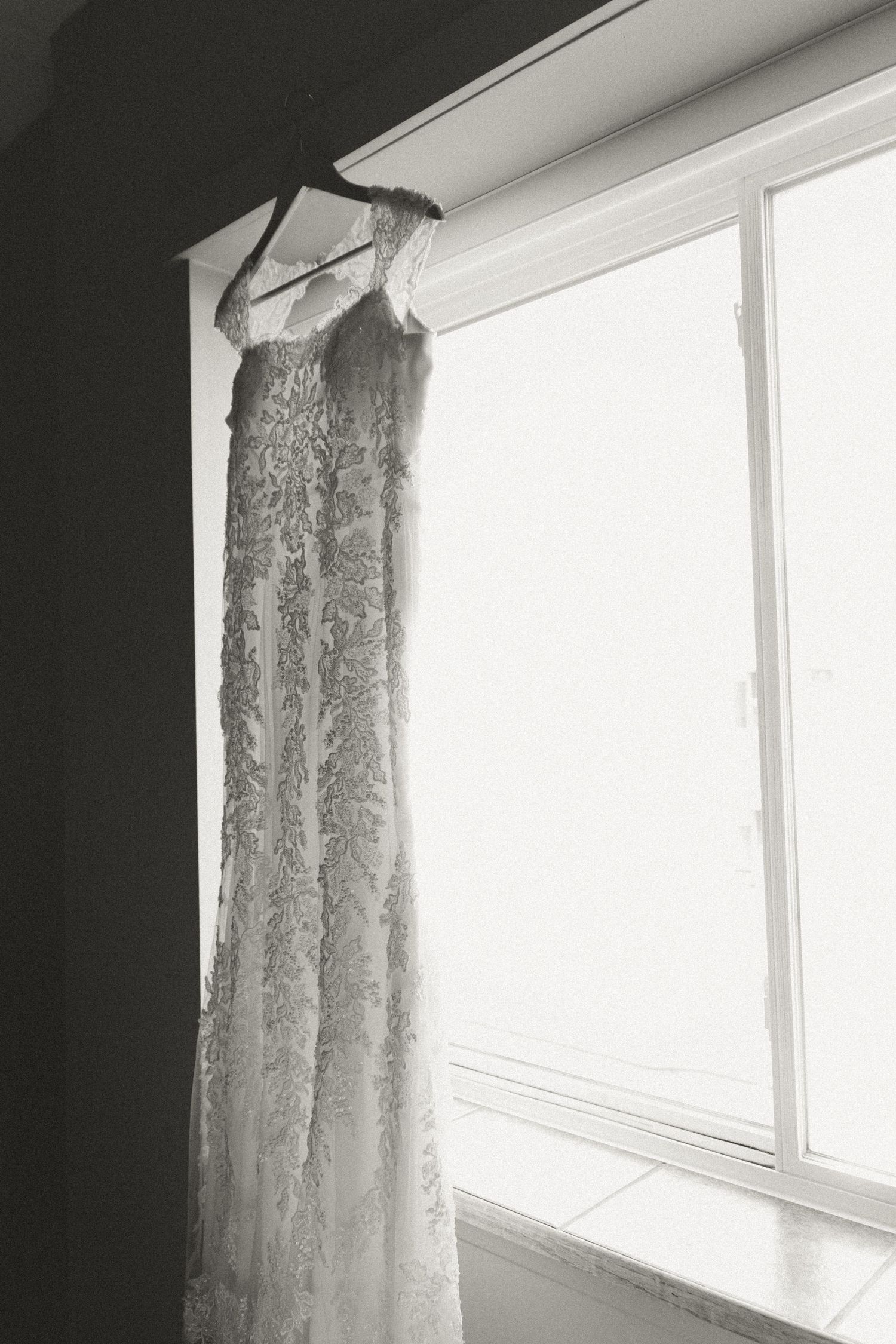 Elegant lace wedding dress hanging by a sunny window in black and white.