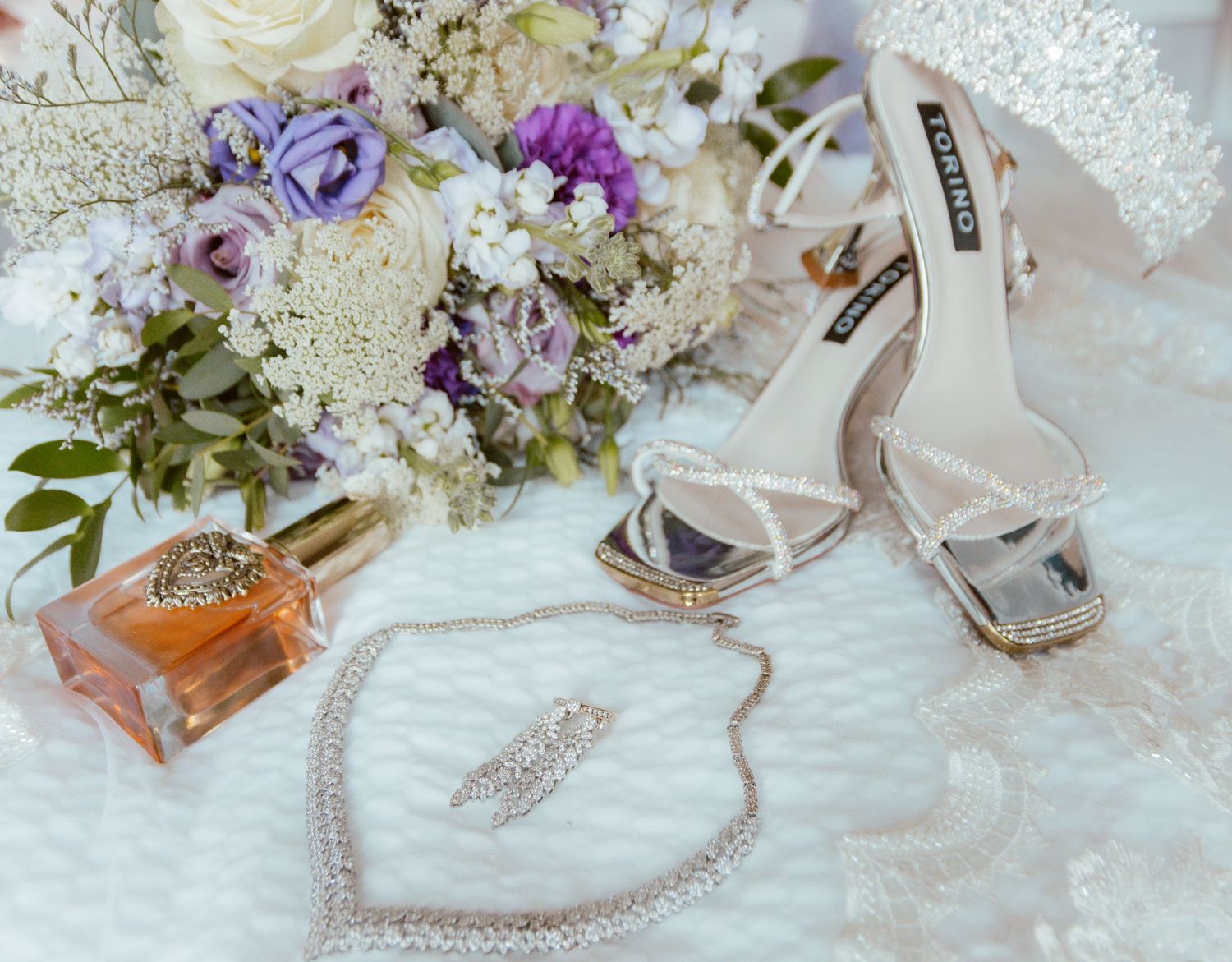 Wedding accessories including flowers, perfume, jewelry and silver heels arranged on lace.