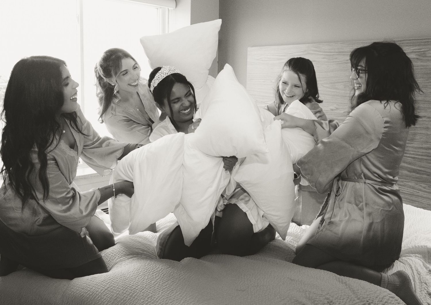 A group of friends laugh and play with pillows during a lighthearted pillow fight in a black and white photograph.