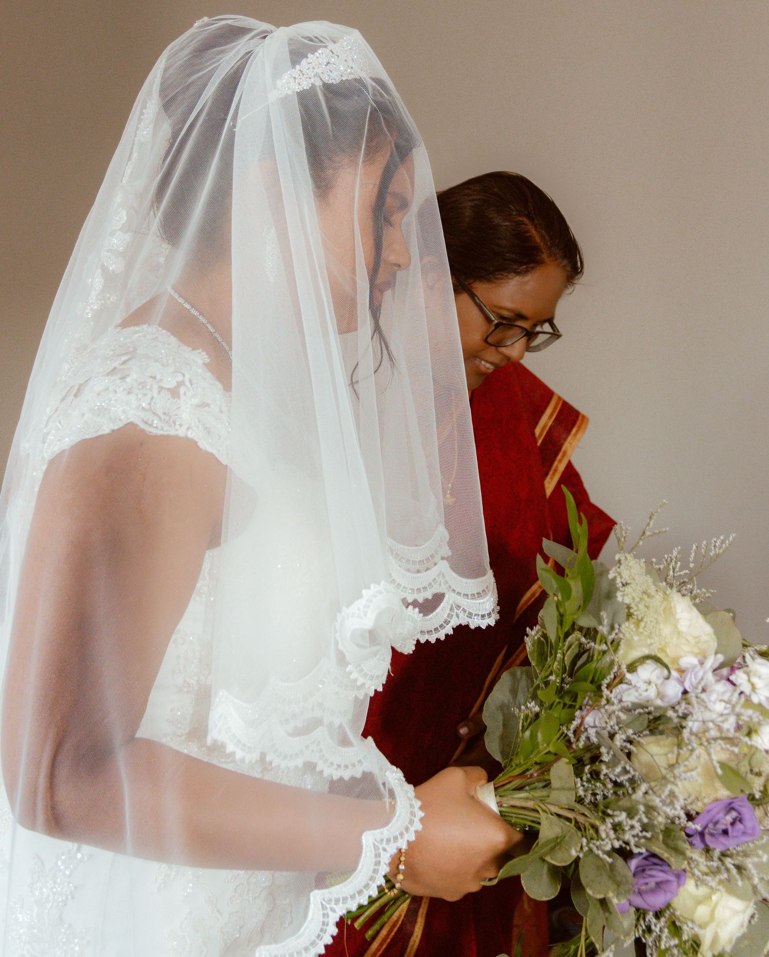 A bride in a lace veil holds a bouquet with purple and white flowers.