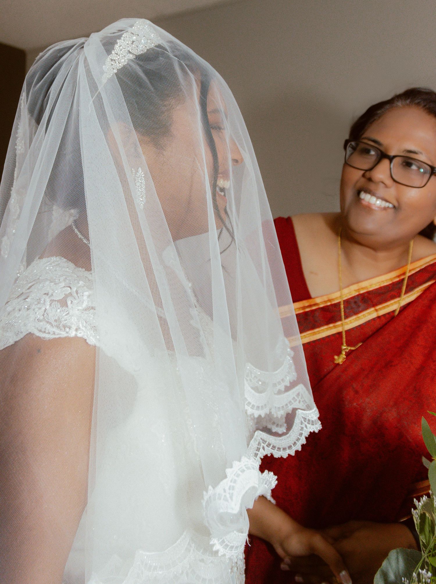 A bride in an elegant lace veil poses with someone wearing a vibrant red traditional sari.