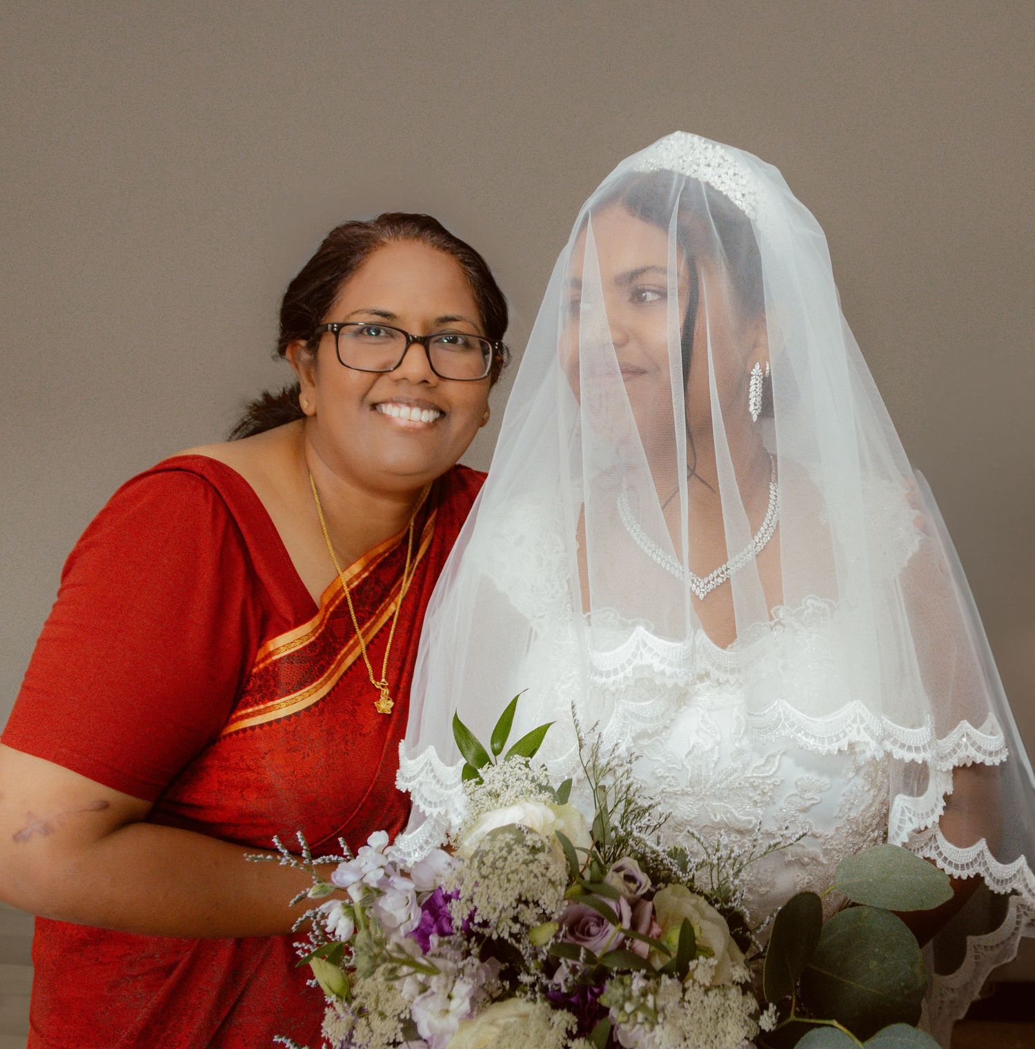A joyful wedding moment between a bride in white veil and someone in a red sari with floral bouquet.