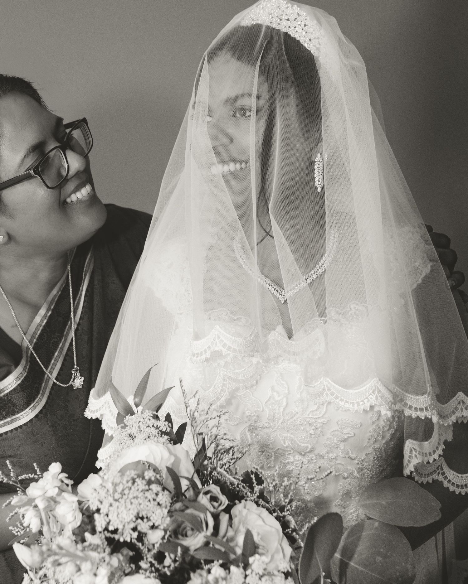 A black and white photo capturing a candid moment between two smiling figures with a bridal veil.