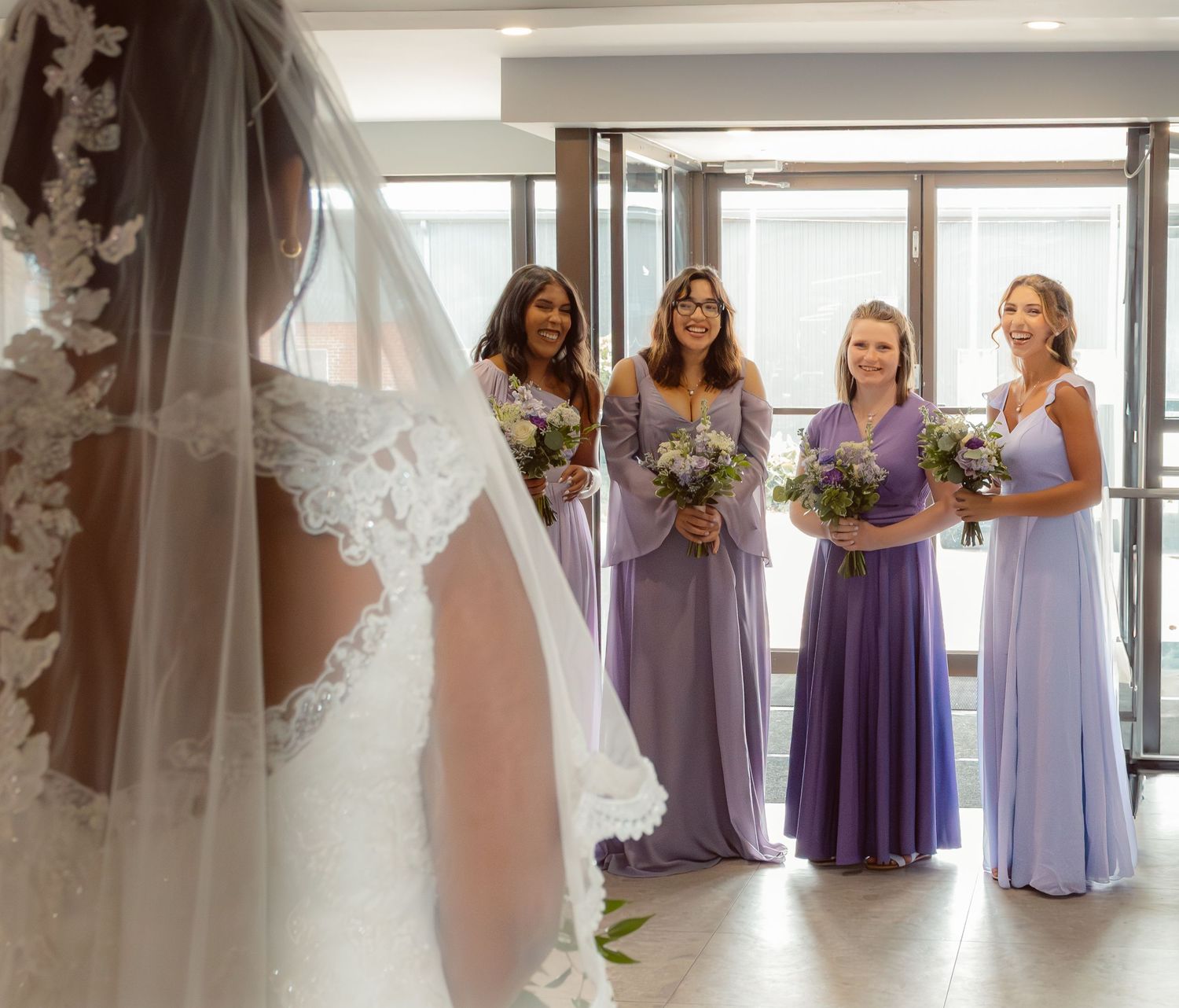 Bridesmaids in purple dresses await the bride in a sunlit room.