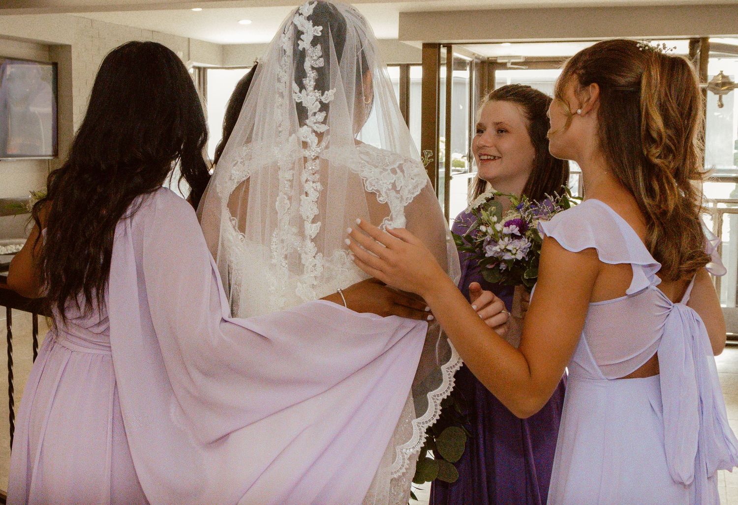 Wedding party members in lavender dresses gather around the bride in her veil.
