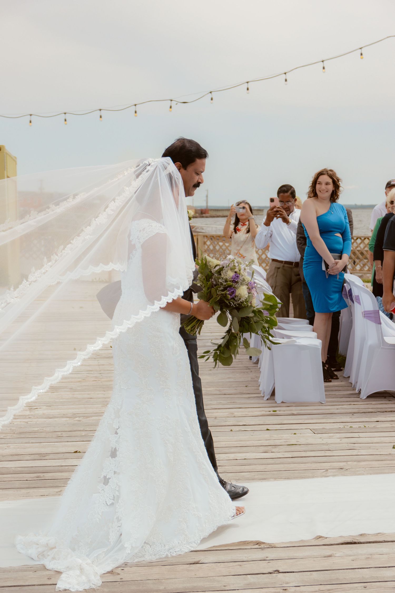 Bride in flowing veil walking along boardwalk at beach wedding ceremony.
