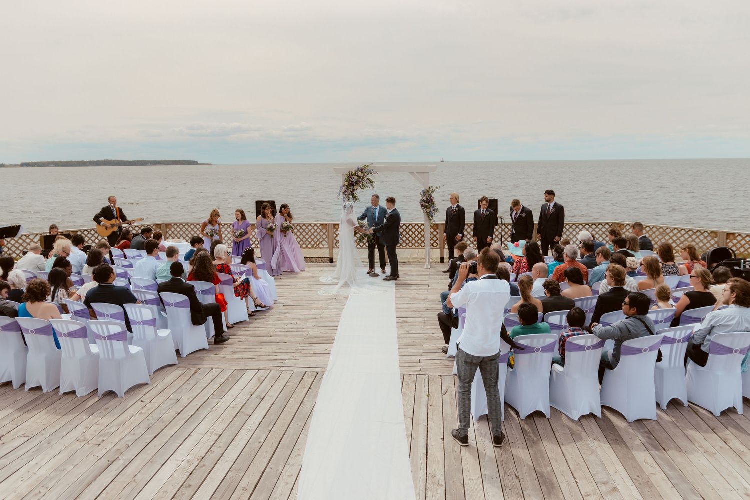 A waterfront wedding ceremony setup with white chairs and guests on a wooden dock.