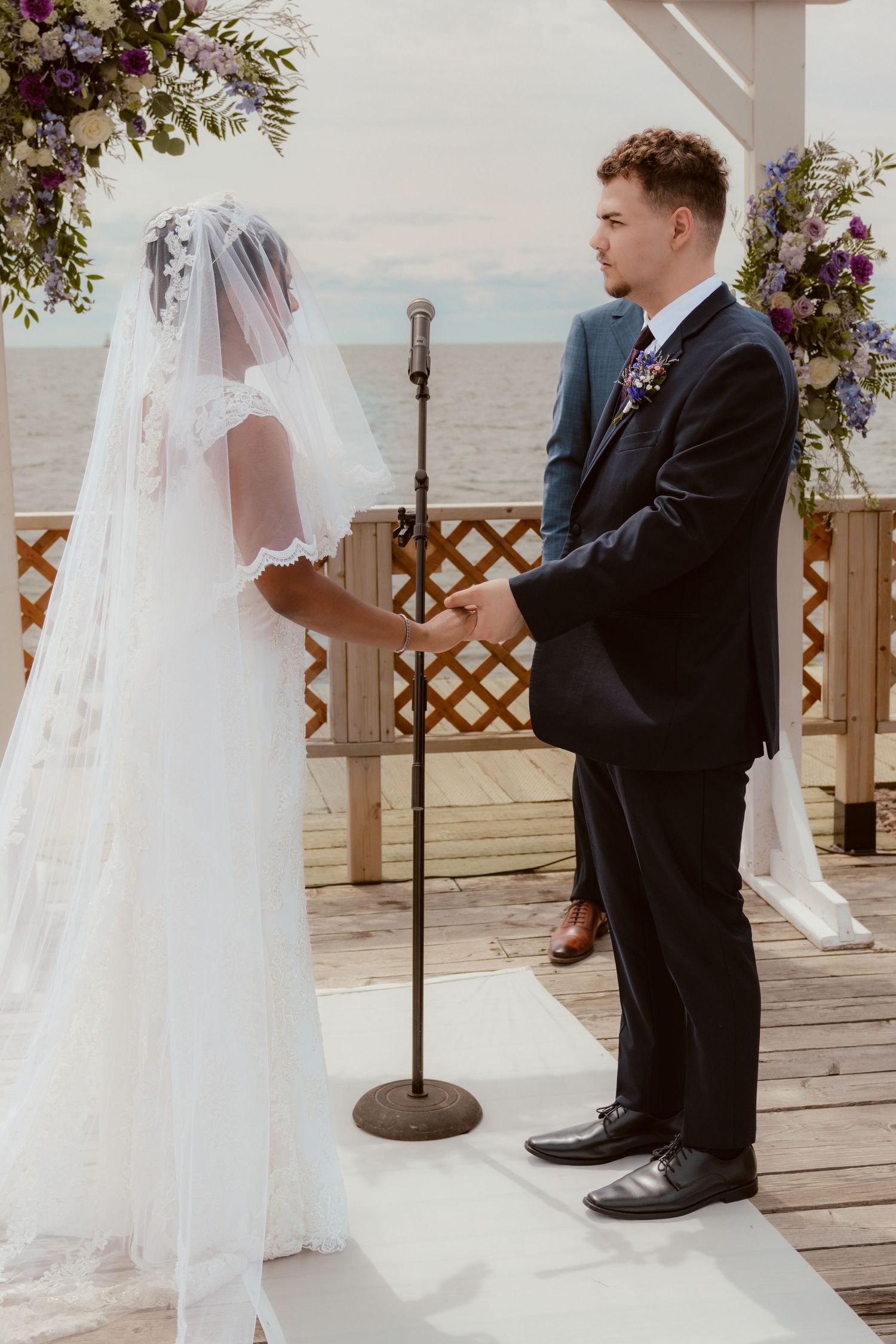 A bride and groom stand at a scenic beach wedding ceremony under a floral arch with a microphone stand between them.