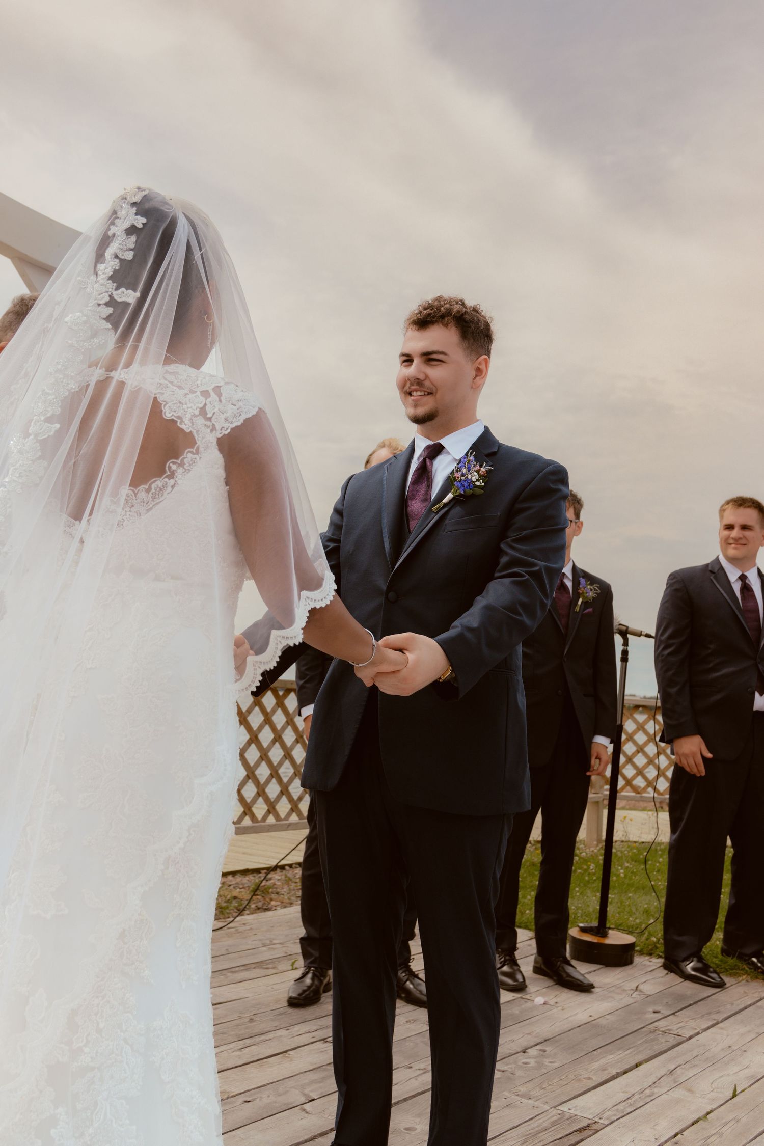 A tender moment during an outdoor wedding ceremony as a couple holds hands on a wooden deck beneath cloudy skies.
