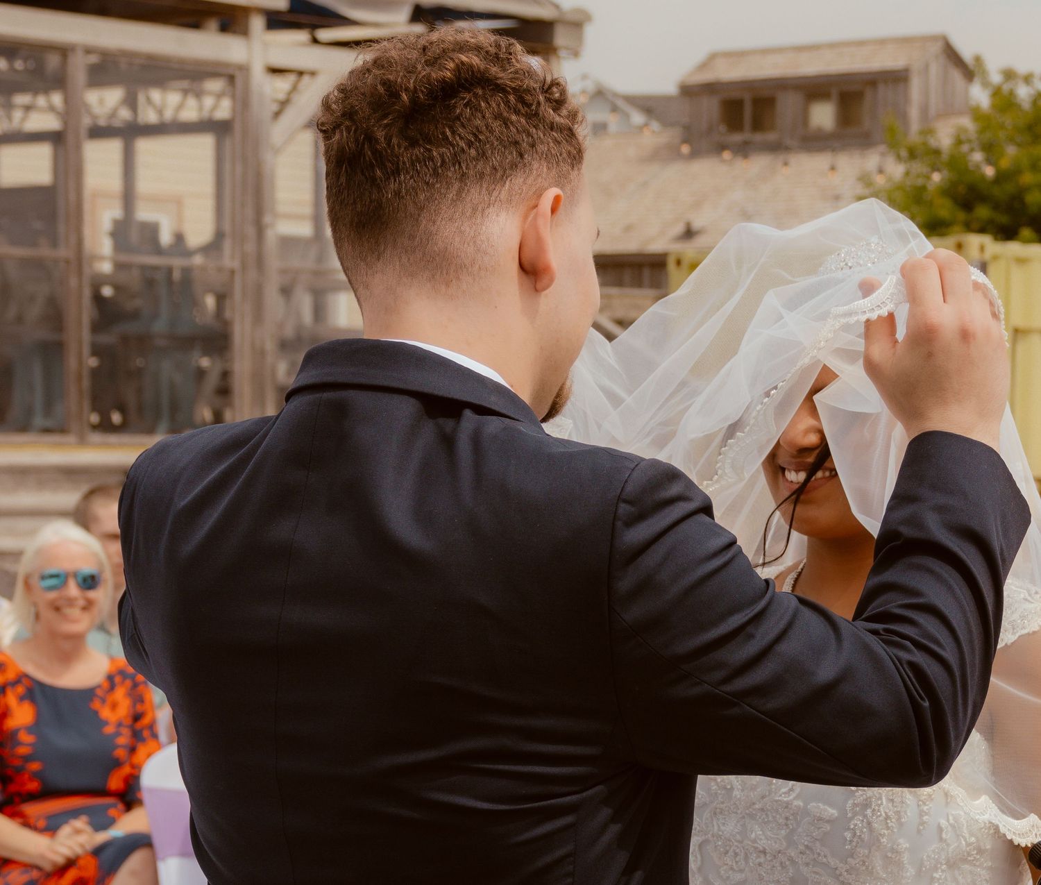 Groom lifts bride's veil during outdoor wedding ceremony as guests watch.