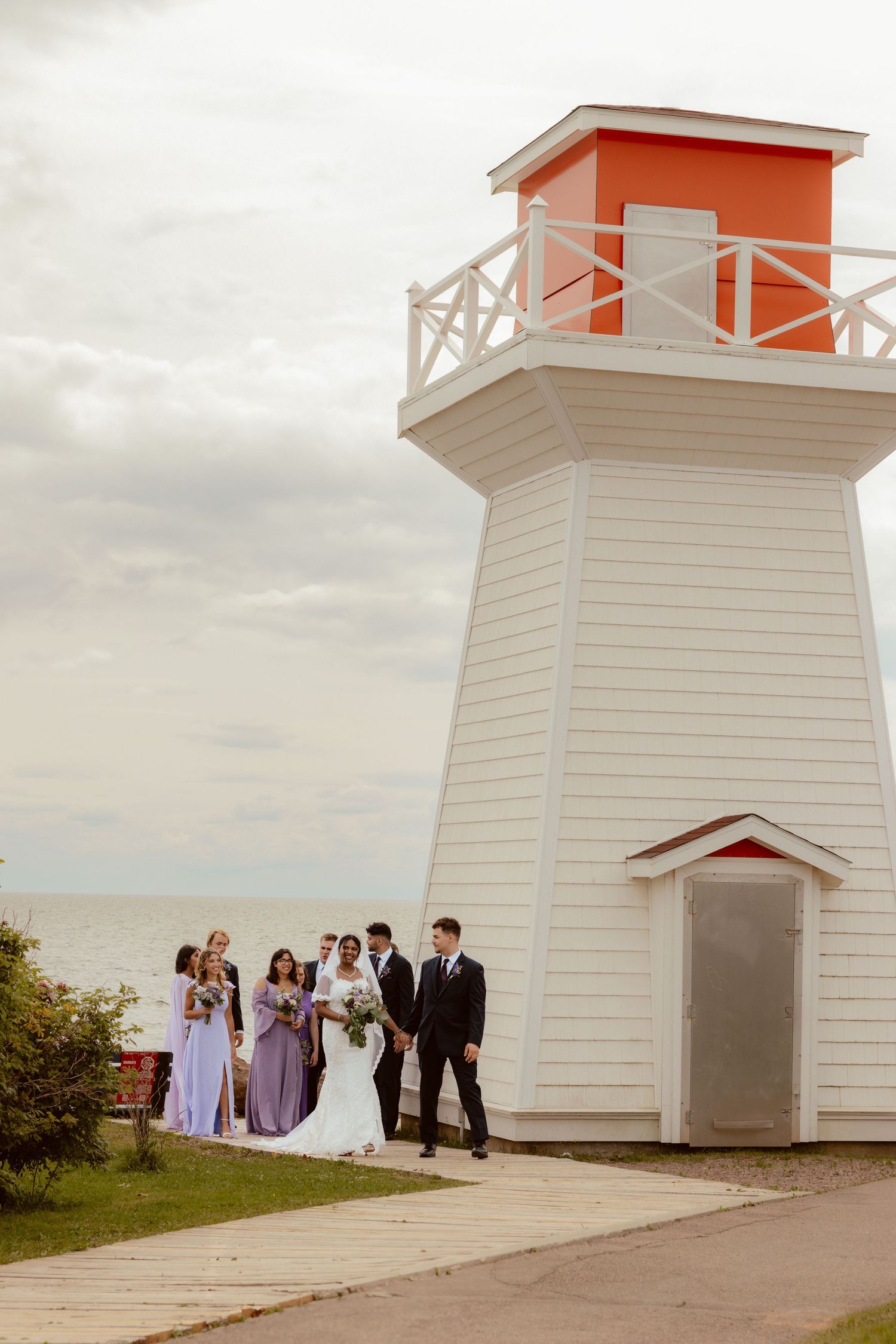 Wedding party poses in front of white and orange lighthouse overlooking the ocean.