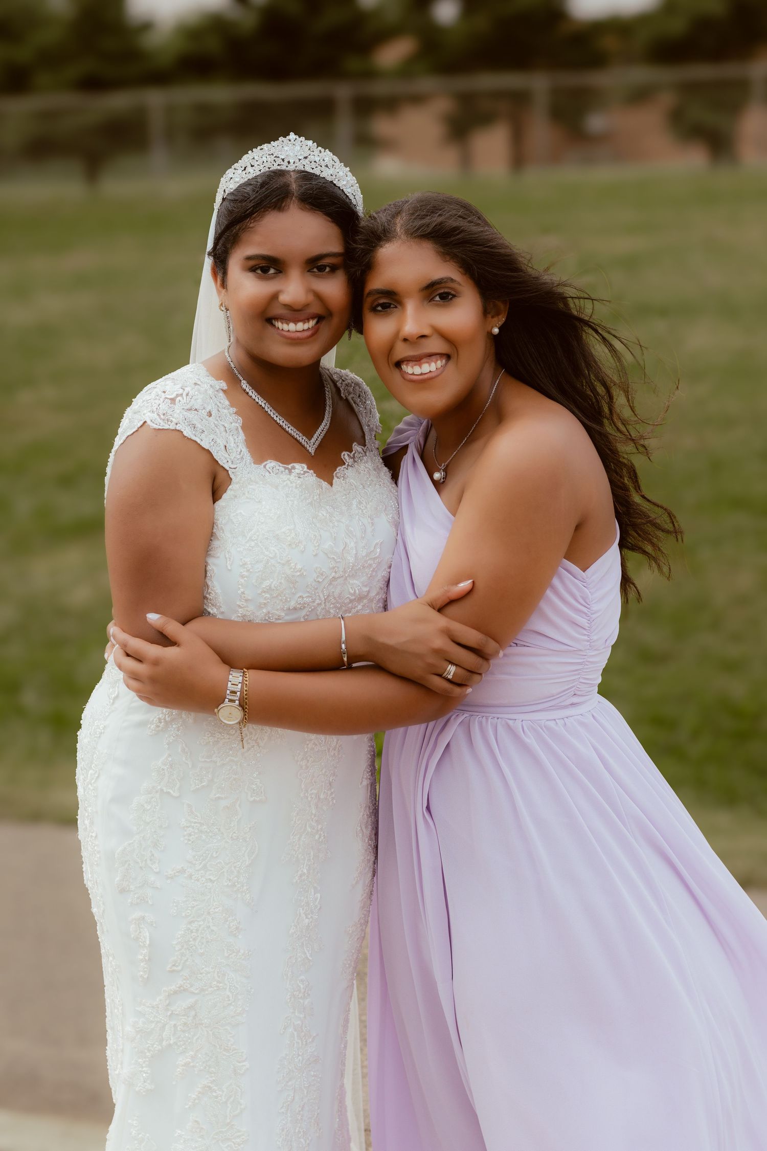Two women in wedding and bridesmaid attire share a joyful embrace outdoors.