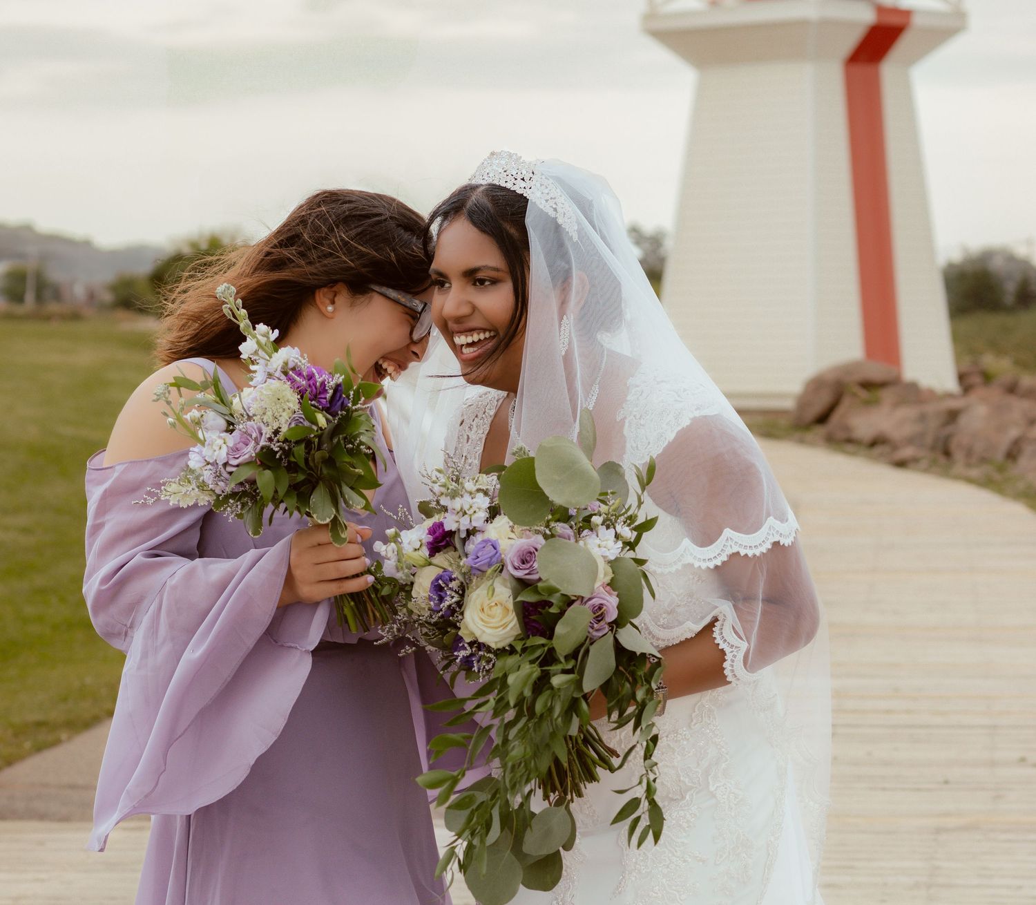Wedding party members share a sweet moment with bouquets near a lighthouse.