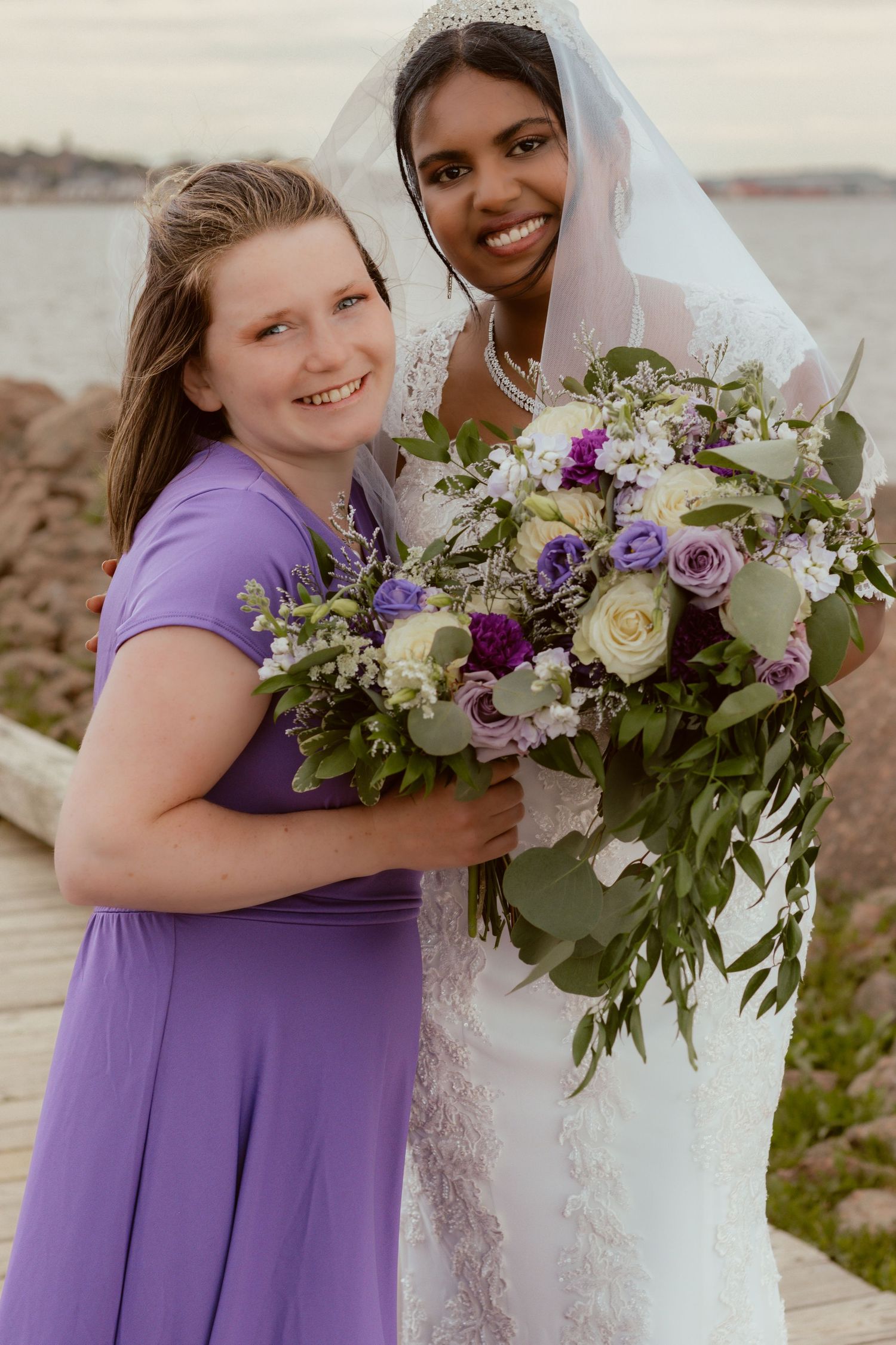 Wedding party members pose with purple and white floral bouquets by the ocean.
