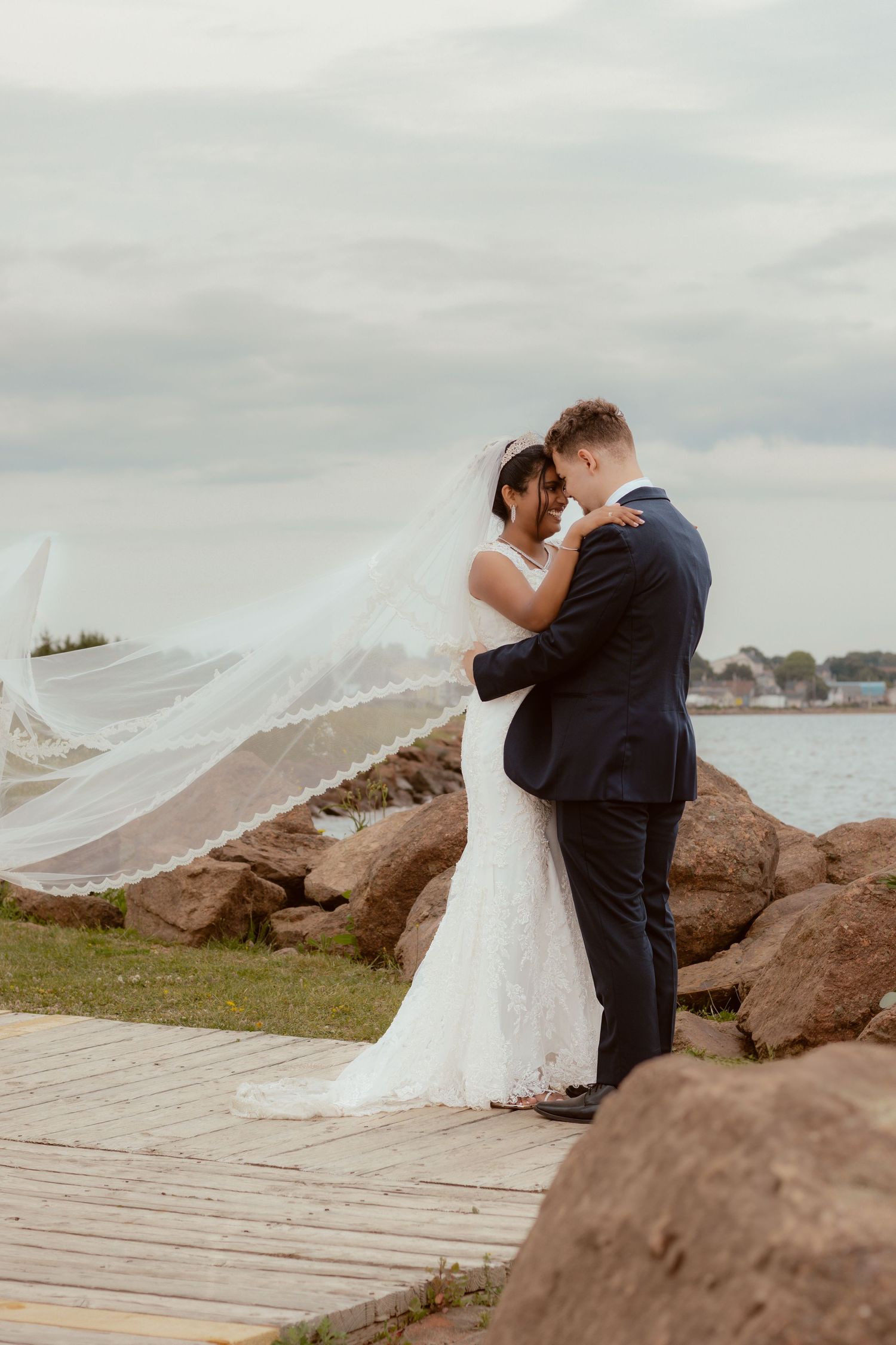 Newlyweds embrace on wooden boardwalk with bride's veil flowing in the wind.