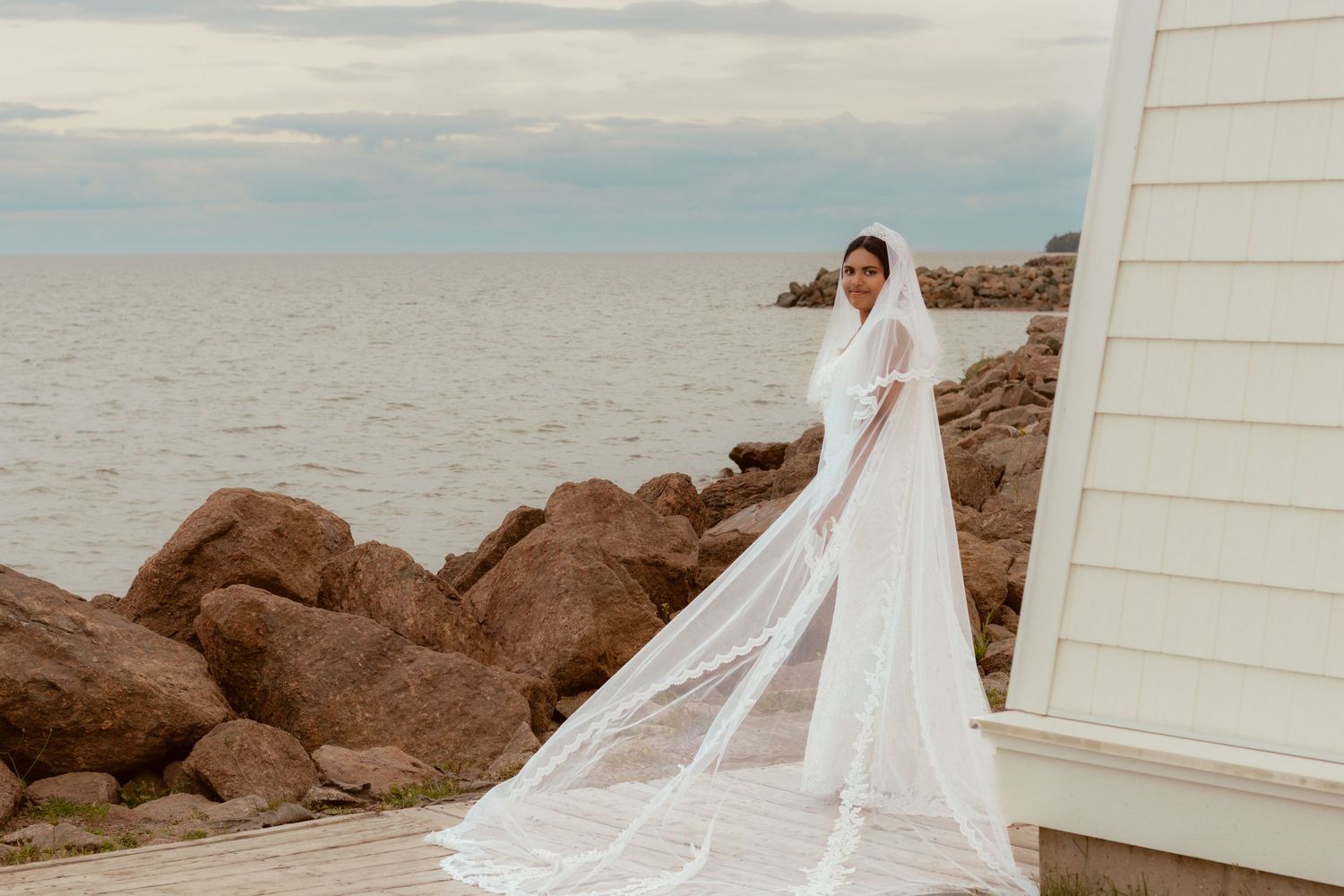 Bride in flowing veil poses by coastal lighthouse and rocky shoreline.