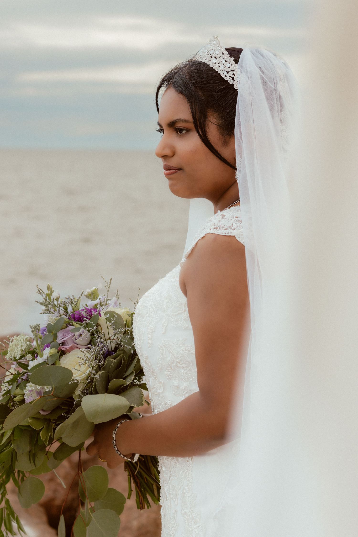 Elegant bride in a lace gown and veil holds a purple and green bouquet against an overcast sky.