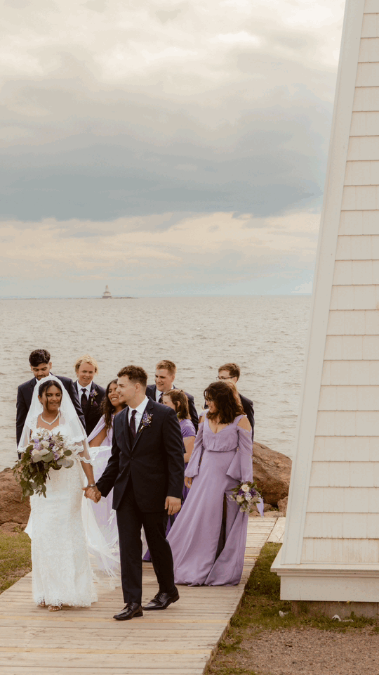 Wedding party walks along wooden boardwalk by lighthouse during coastal celebration.