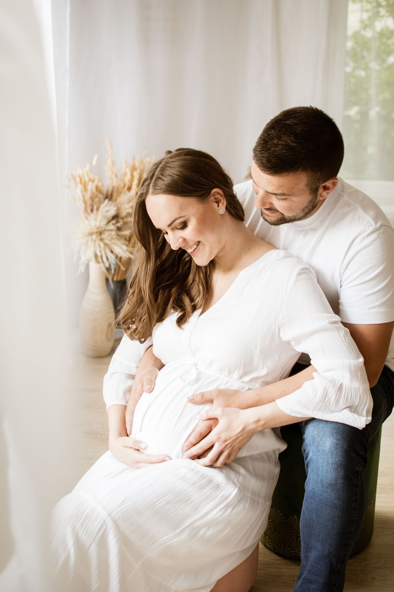 Tender maternity portrait of couple in white by window with dried flowers.