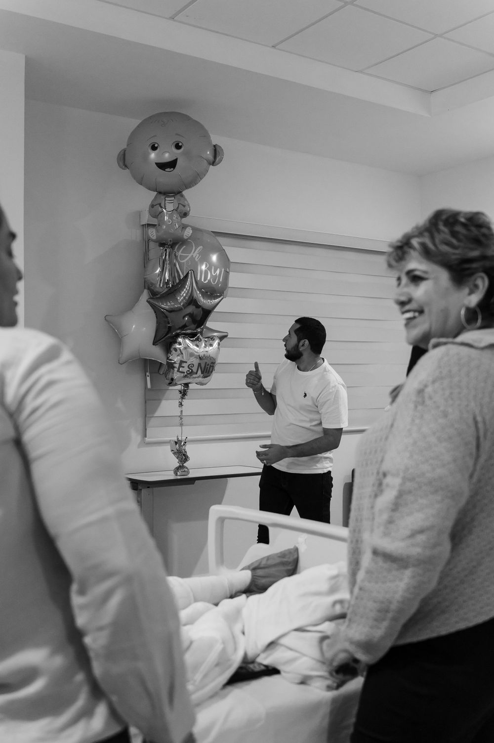 Hospital room decorated with balloons as visitors gather around patient bed.
