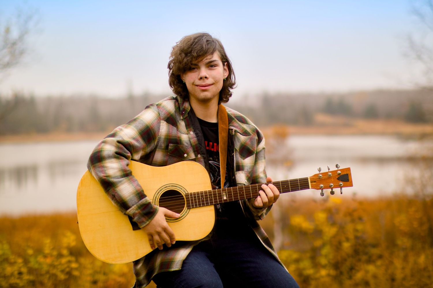A person in plaid flannel plays acoustic guitar outdoors by a lake during autumn.
