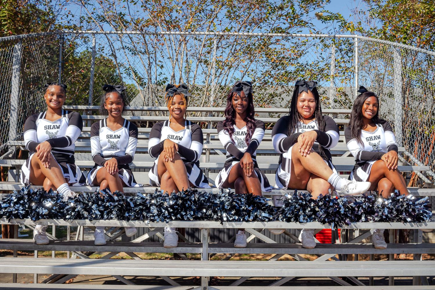 Cheerleaders in matching blue and white uniforms pose together on metal bleachers during a sunny autumn day.