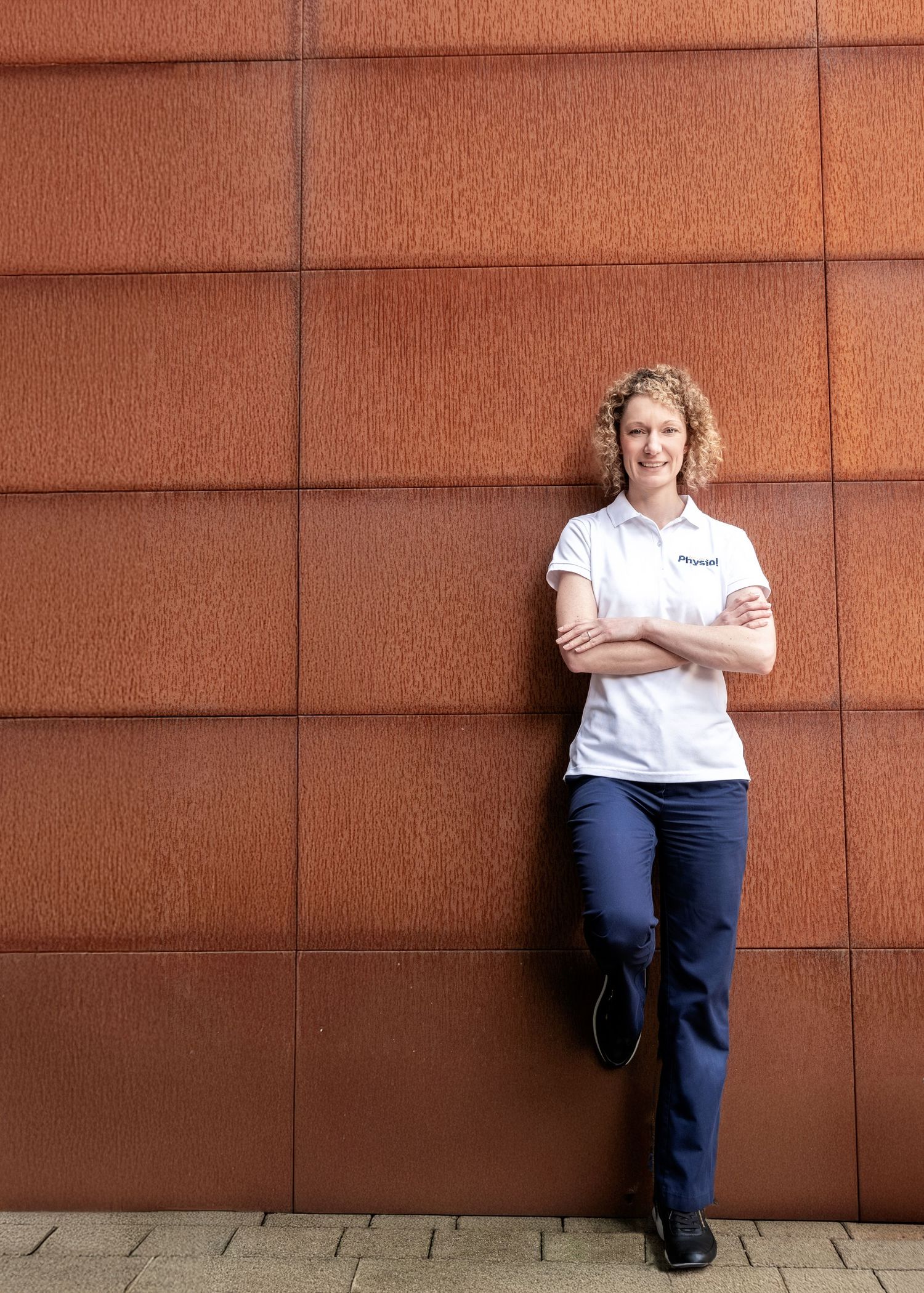 Professional portrait of a person in a white polo shirt and navy pants standing against a textured rust-colored wall.