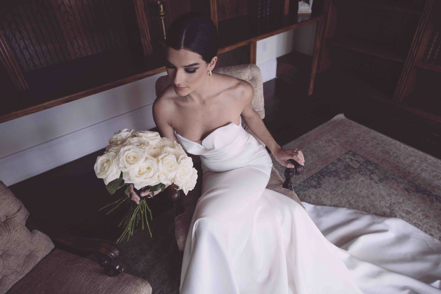 Bride in white strapless wedding gown holds bouquet of white roses while seated.
