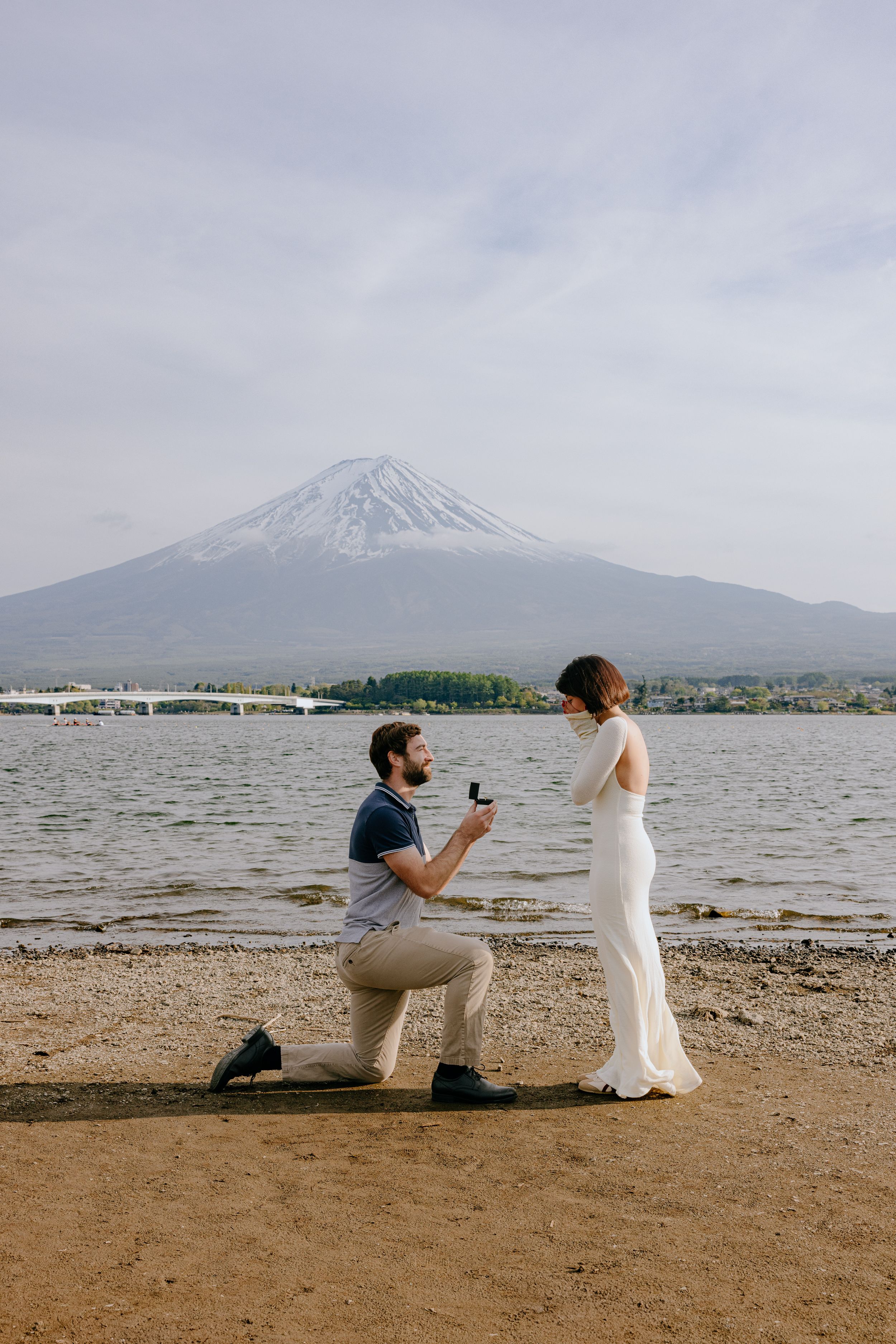 A romantic proposal scene unfolds on a beach with Mount Fuji's snow-capped peak visible in the background.