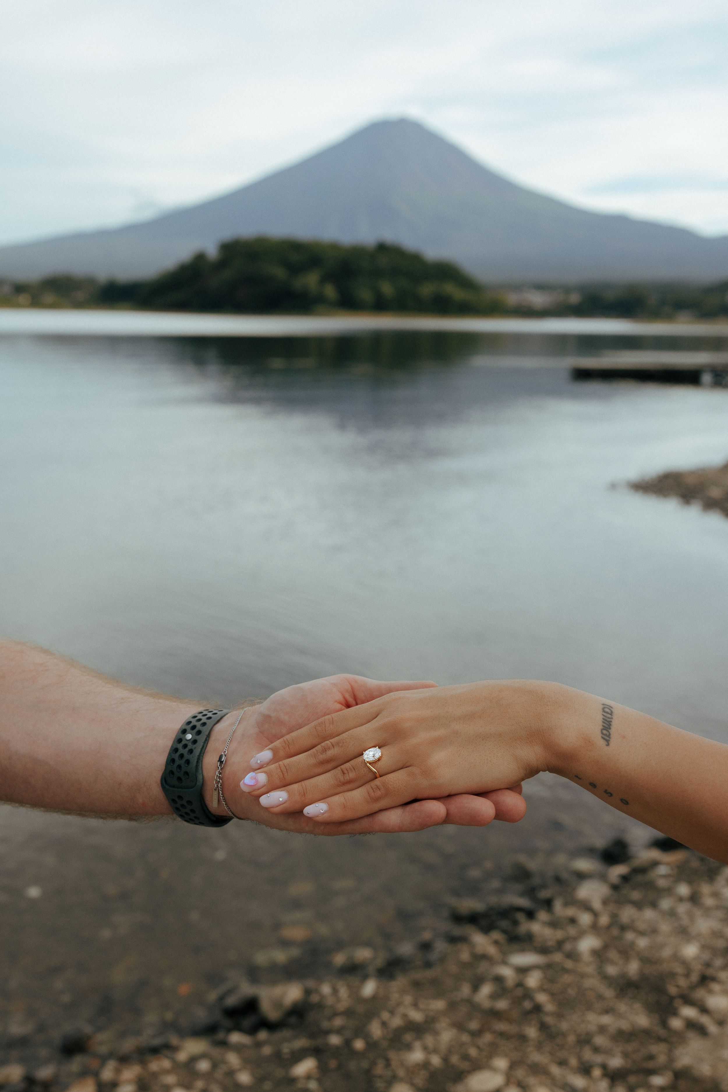 Two hands reaching out with an engagement ring against the backdrop of Mount Fuji and a serene lake.