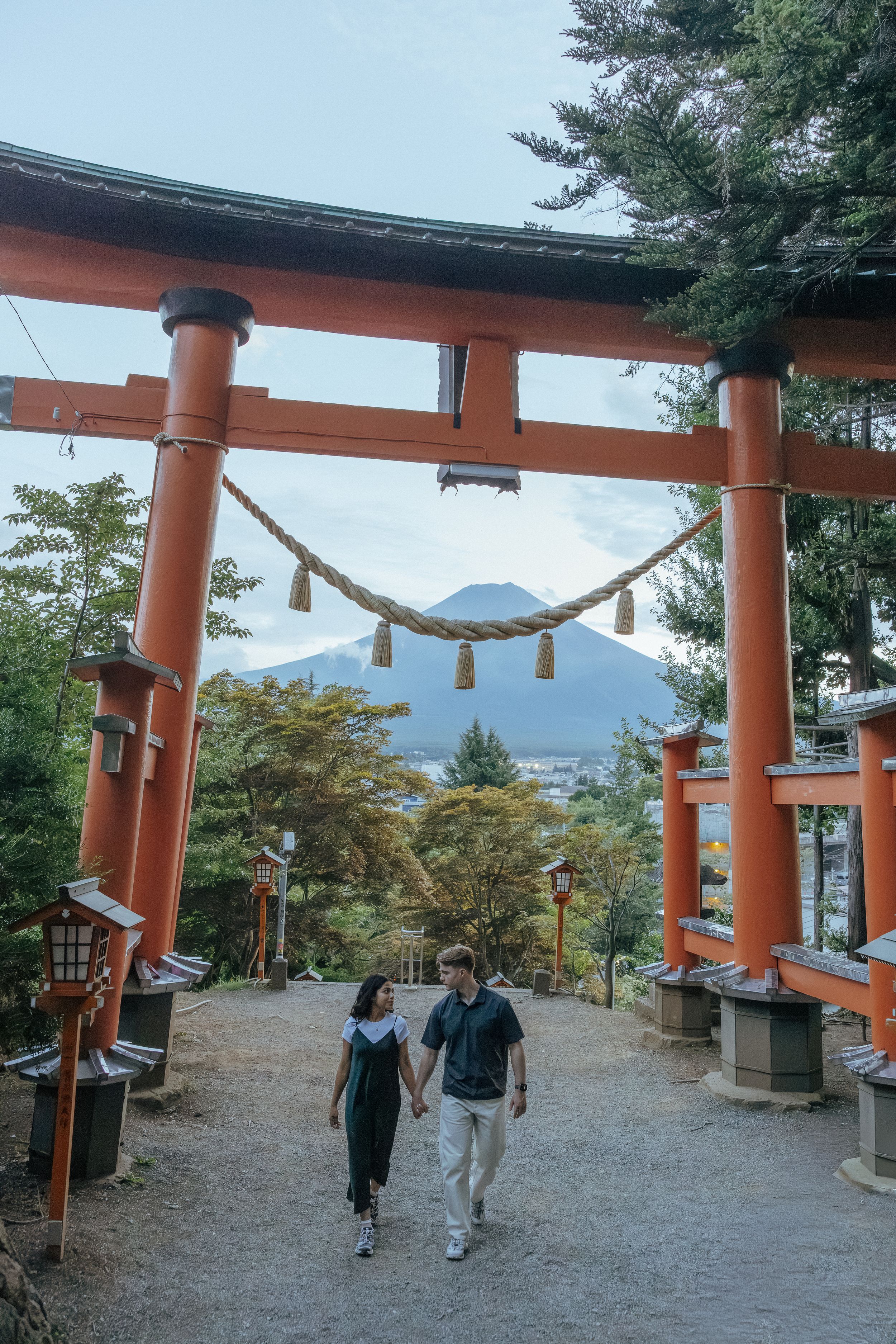 Orange torii gates line a pathway at a traditional Japanese shrine surrounded by trees on a cloudy day.