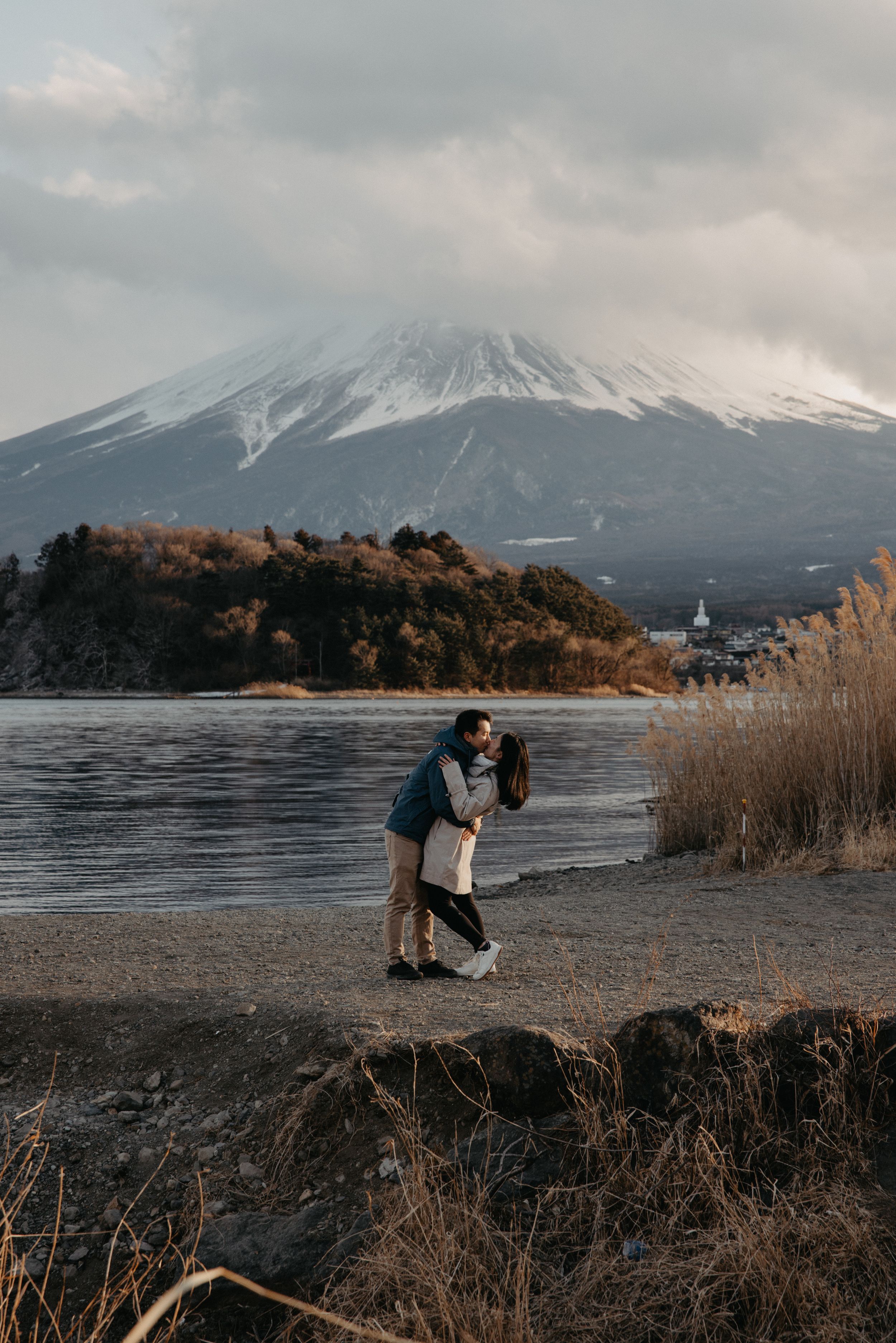 A couple embraces by a lake with Mount Fuji rising majestically in the background during winter.