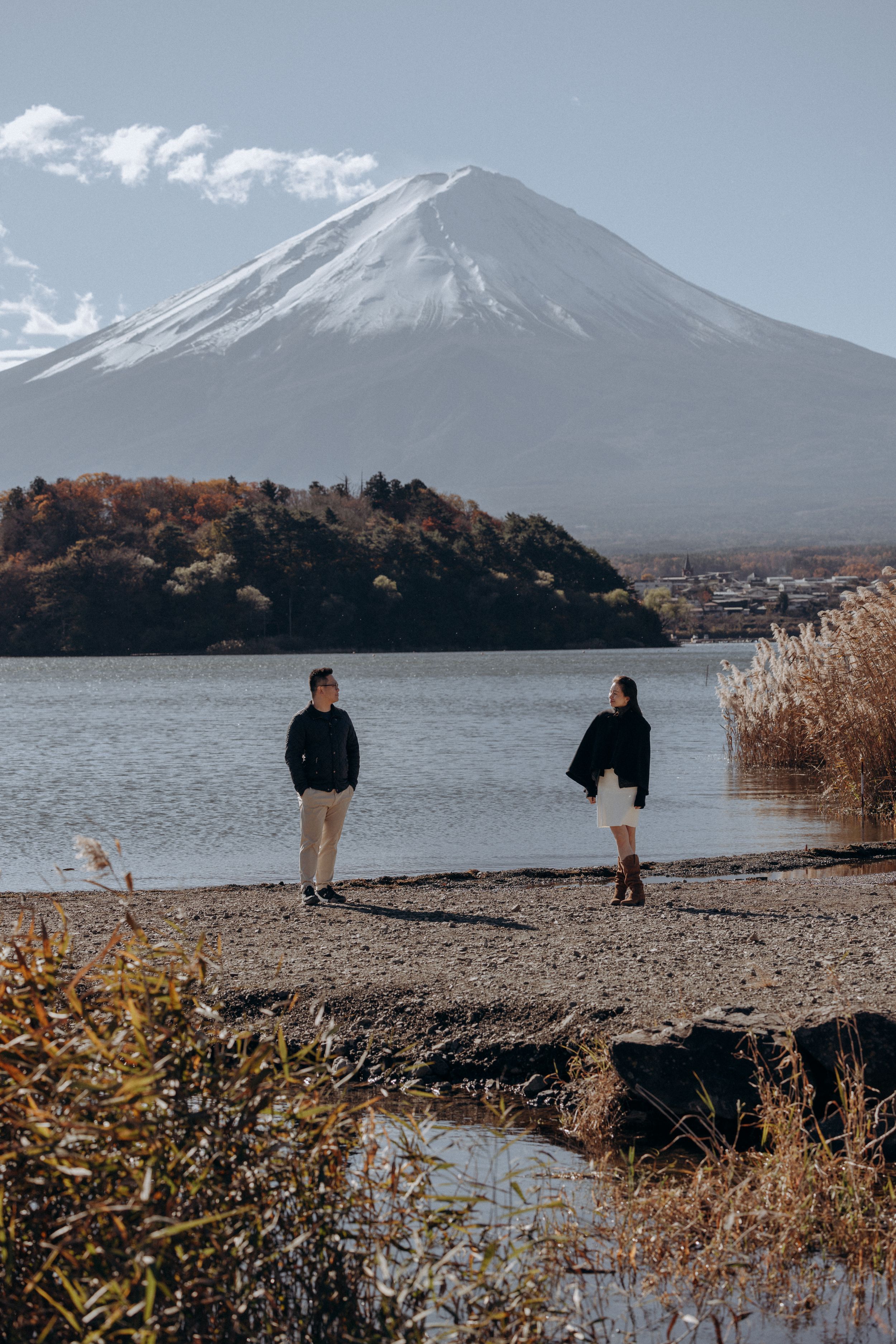 Two people stand by Lake Kawaguchi on a rocky shore with Mount Fuji's snow-capped peak rising majestically in the background.