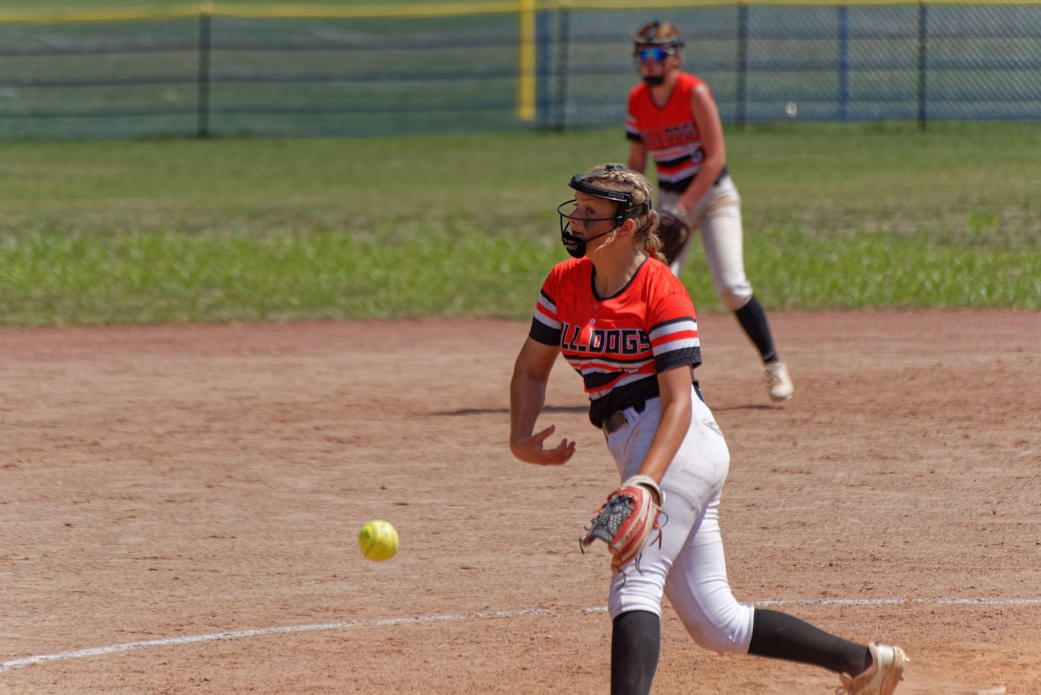 Female softball player in uniform pitching yellow ball during game on dirt field.