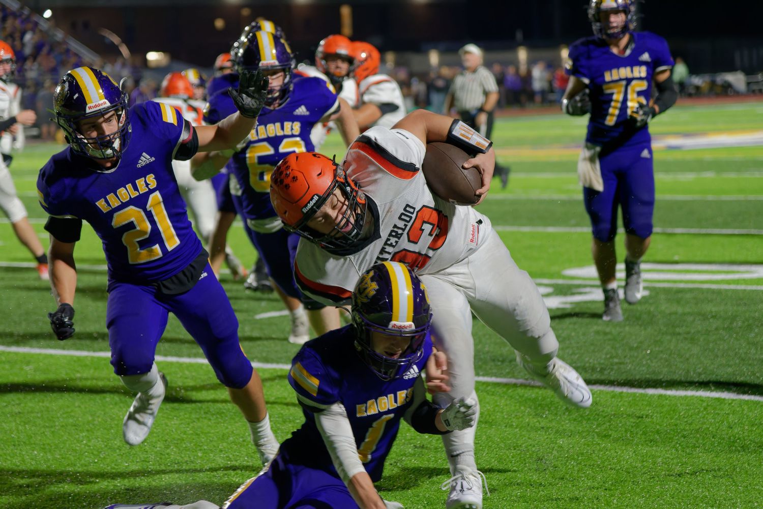 High school football players collide during a tackle as a running back pushes through defenders in purple jerseys.