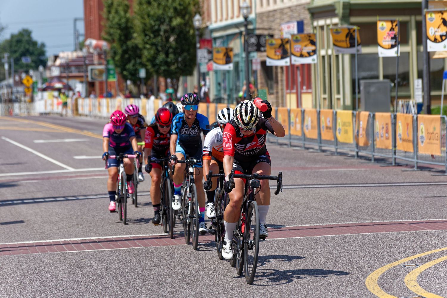Cyclists race in a line formation along an urban street during a competitive cycling event on a sunny day.