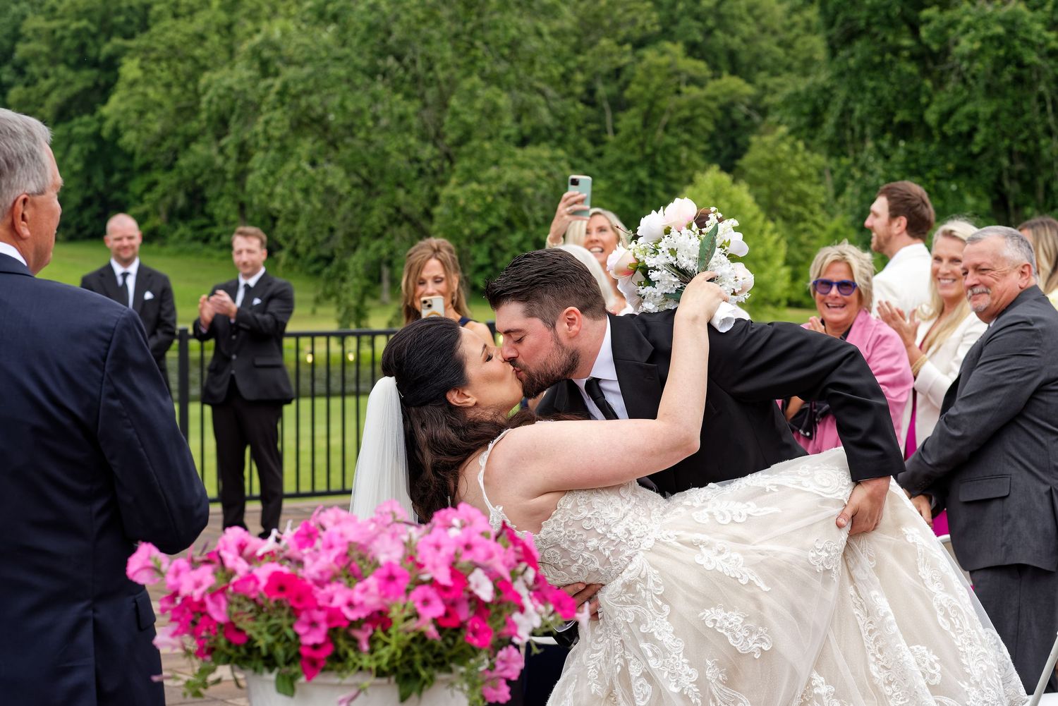 A group of wedding guests gathered outdoors in formal attire with bright pink floral decorations in the background.