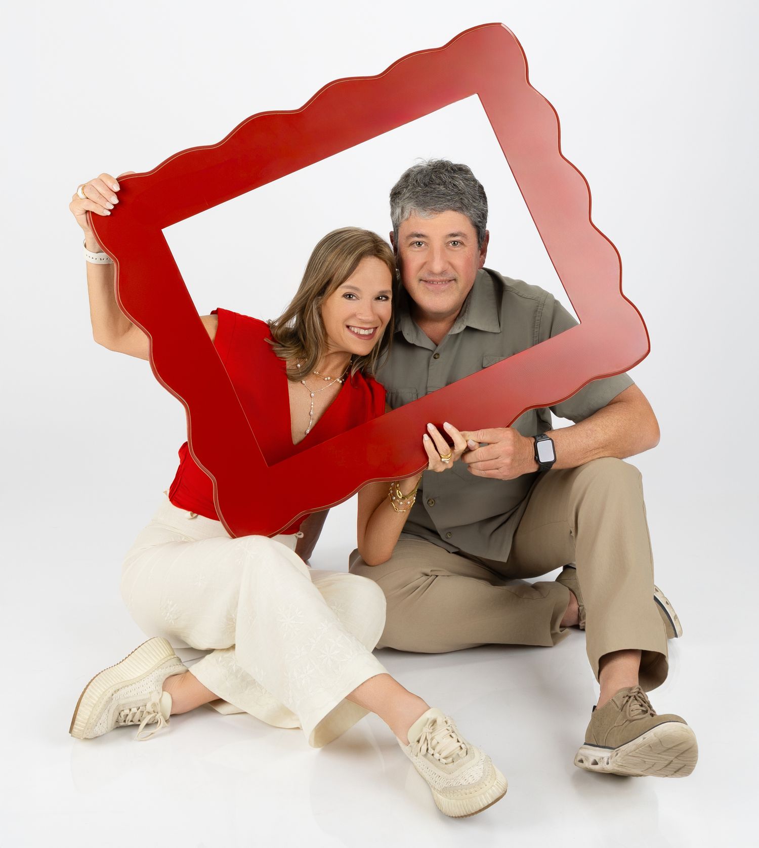 Couple poses playfully with large red decorative frame against white studio background.