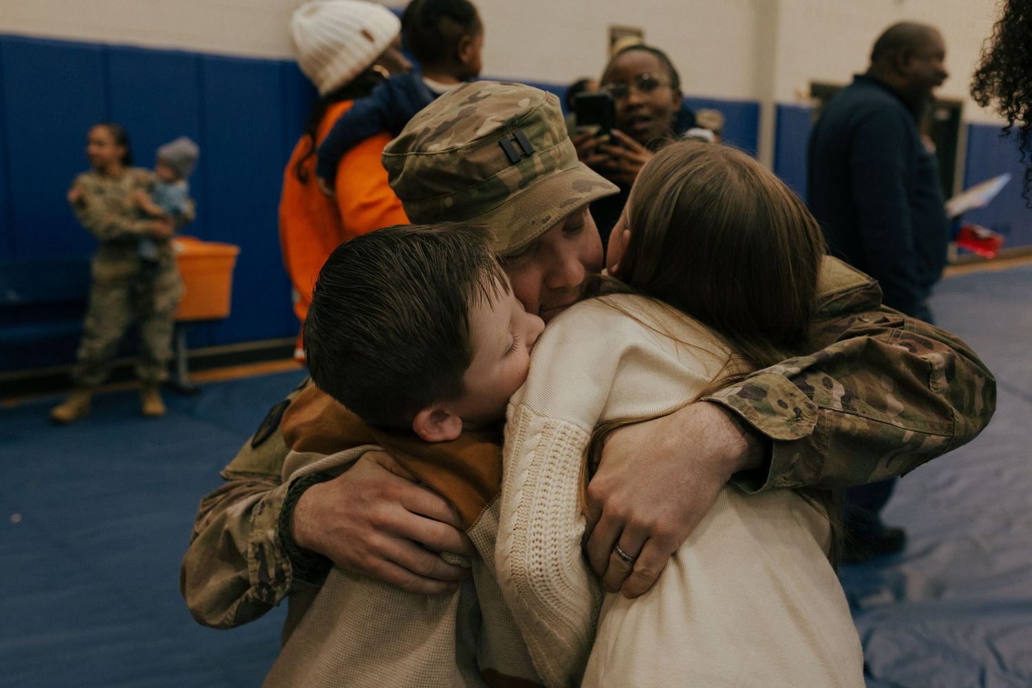 Military service member in camouflage uniform embraces loved one during emotional homecoming reunion at gymnasium.