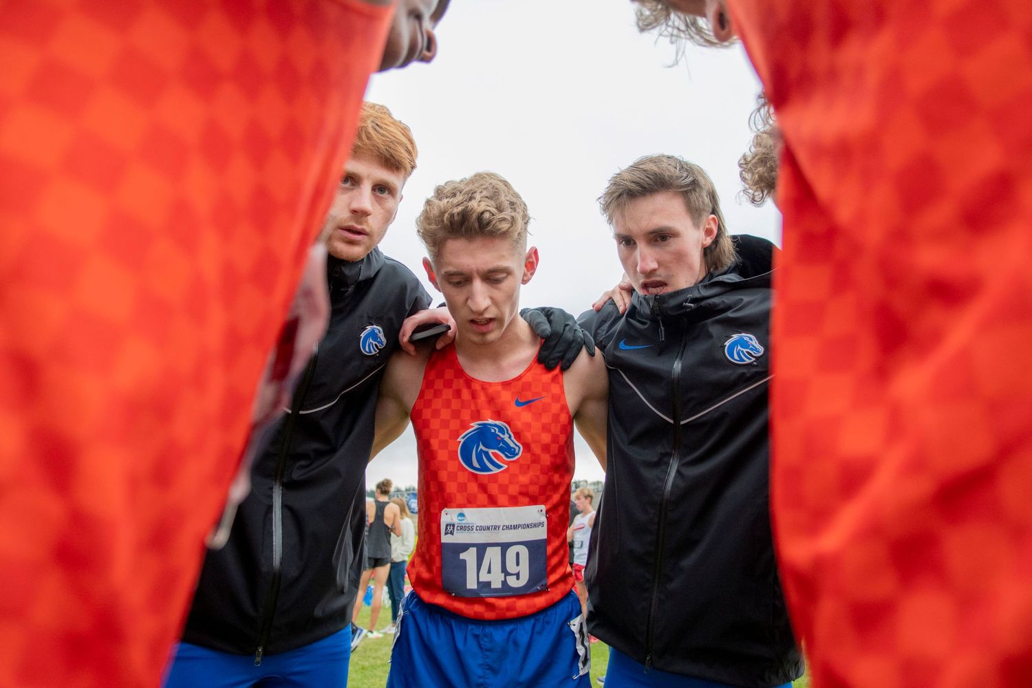 Three athletes in orange jersey and black jackets huddle together during a cross country race event.