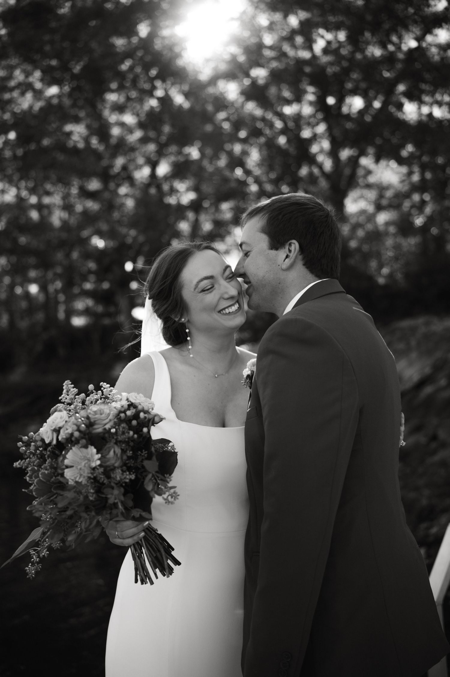 A black and white wedding photo capturing a romantic moment between a bride with a bouquet and groom outdoors.