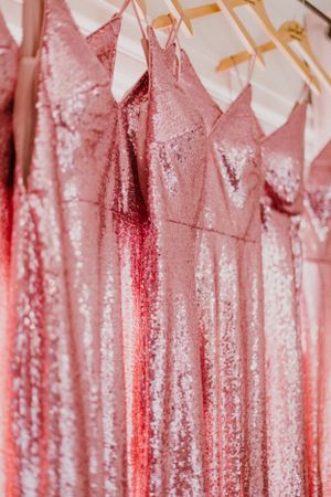 Row of sparkly pink sequined bridesmaid dresses hanging on wooden hangers against a white background.