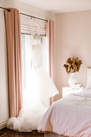 Elegant white wedding dress hanging by window with sheer pink curtains and white bedding.