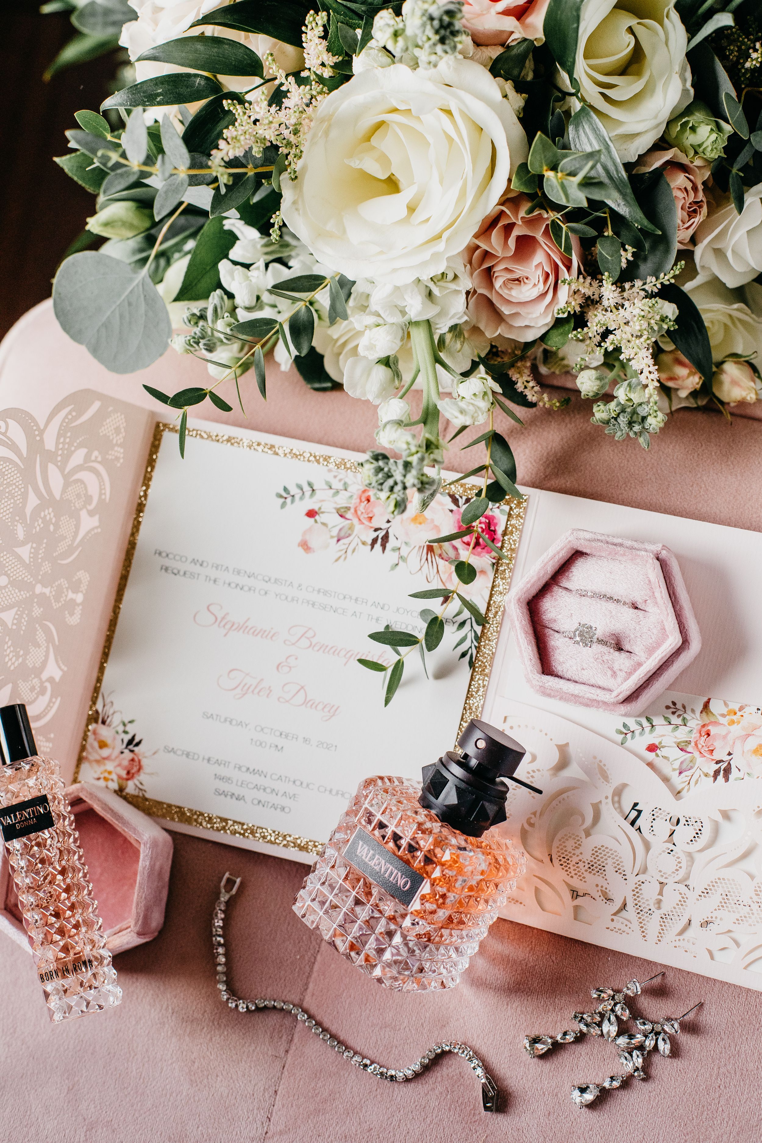 Wedding details flatlay with white roses, invitation, and accessories on pink fabric.