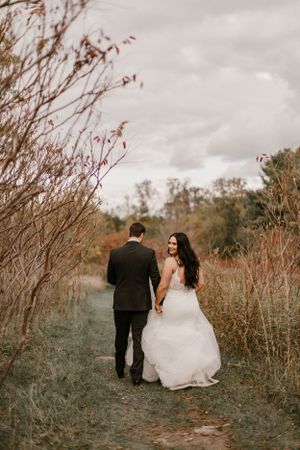 Couple walking down nature path holding hands, bride in white gown and groom in black suit.